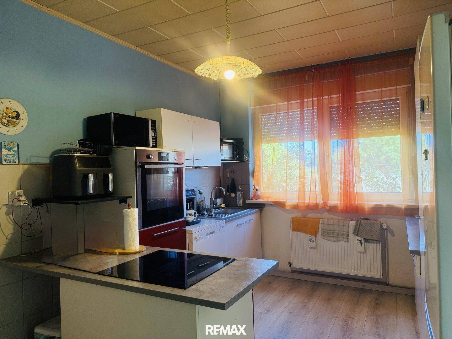 Helle Küche mit blauen Wänden, weißen Einbauschränken und einem Induktionskochfeld. Das Fenster sorgt für viel Tageslicht.