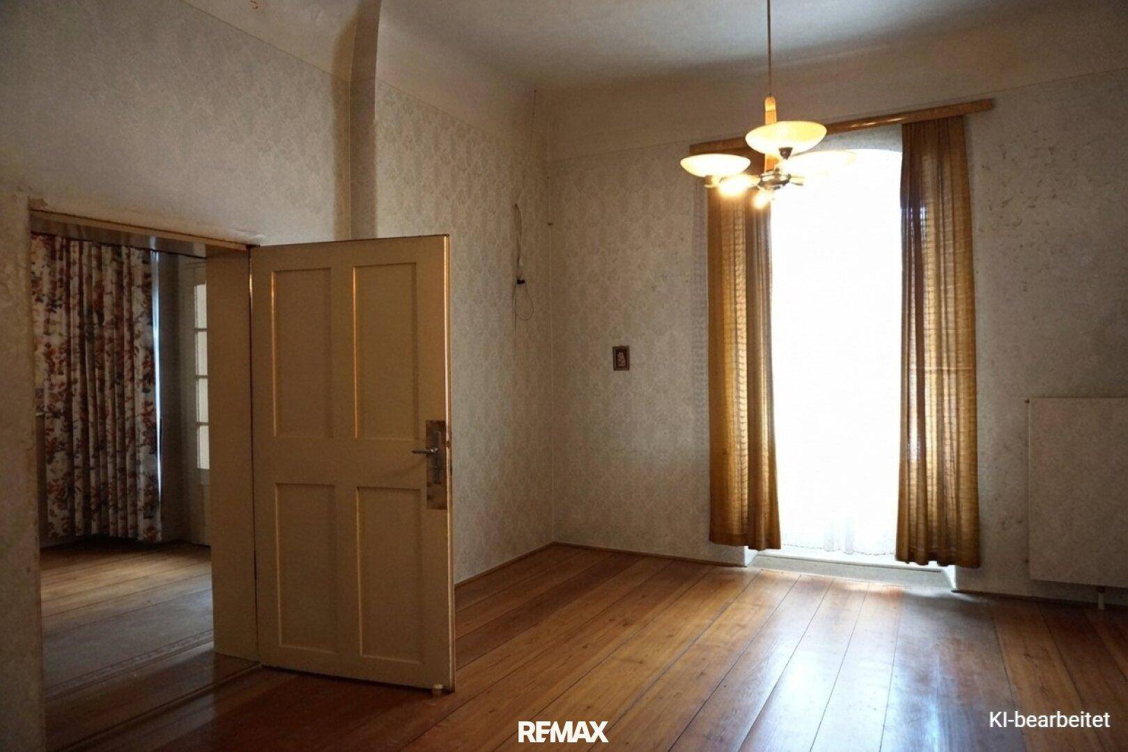 Zimmer mit gemusterter Tapete, Holzdielenboden und einem hohen Fenster, das auf eine Renovierung wartet.