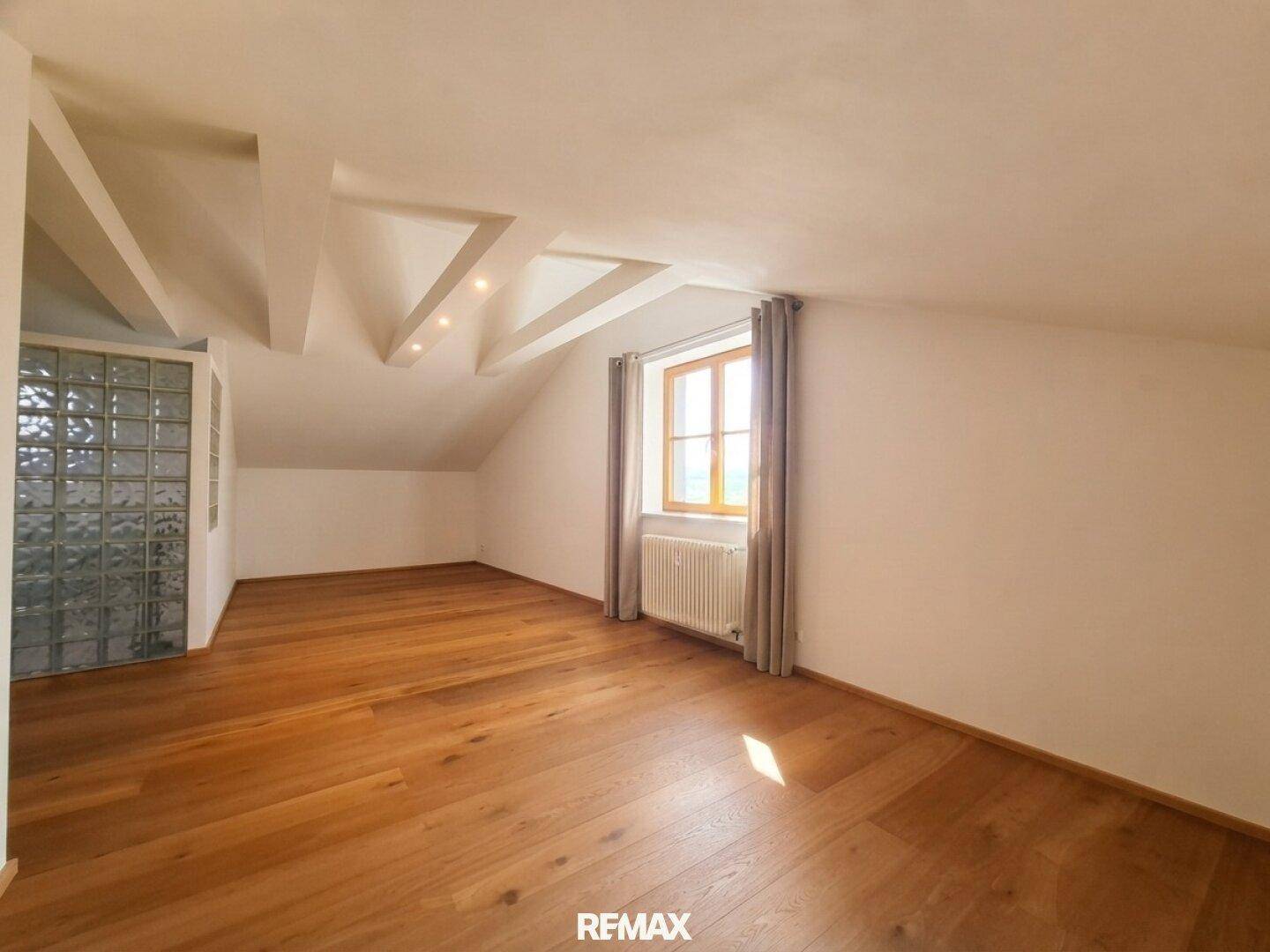 Helles Zimmer im Dachgeschoss mit Holzboden, sichtbaren Balken und Fenster.