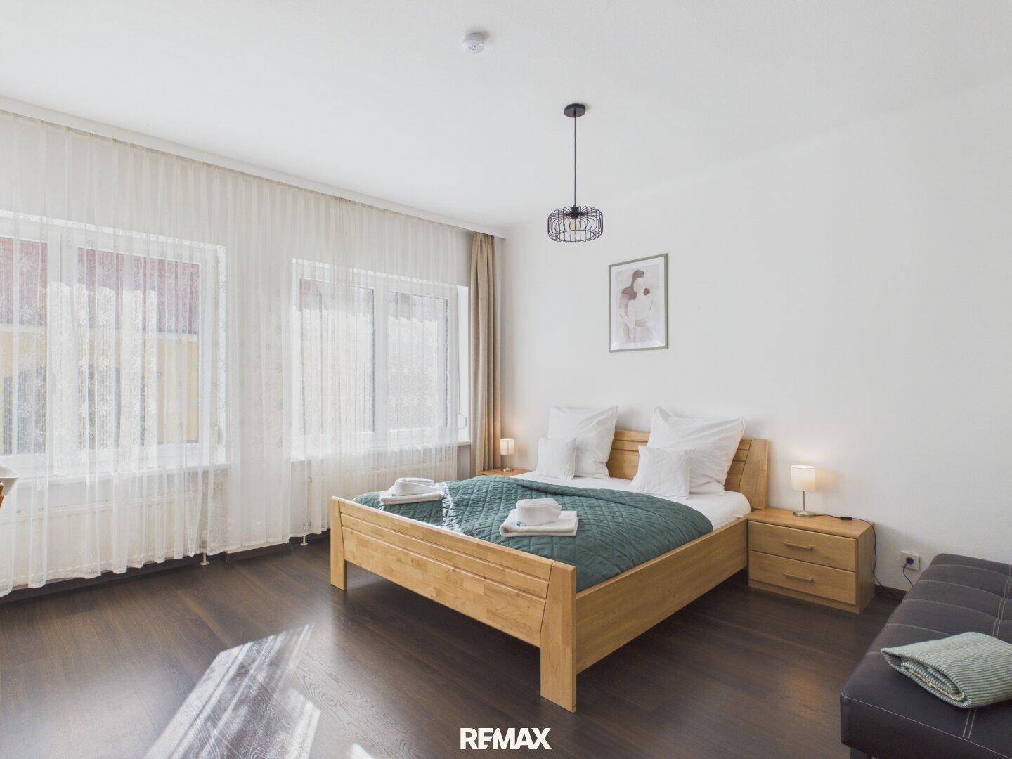 Gemütliches Schlafzimmer mit Holzbett, dunklem Holzboden und hellen Vorhängen an den Fenstern.
