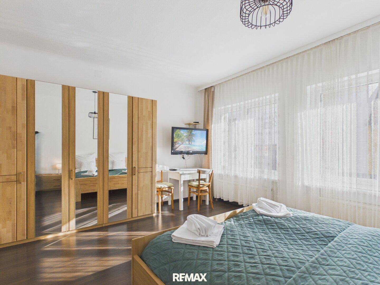 Schlafzimmer mit großem Spiegelschrank, dunklem Holzboden und einem kleinen Arbeitsbereich am Fenster.
