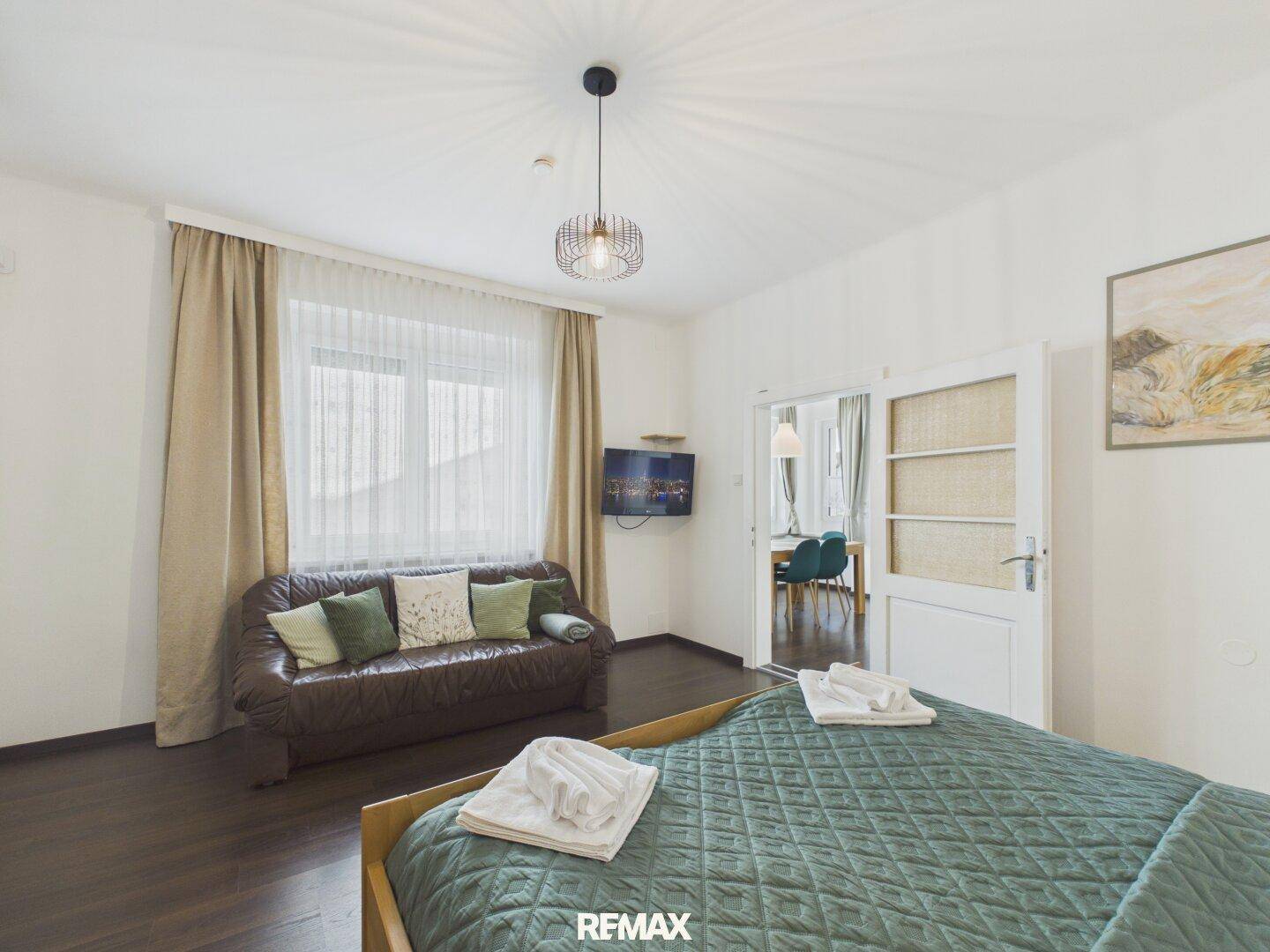 Schlafzimmer mit Bett und Sofa, das durch eine offene Tür zum Essbereich führt.