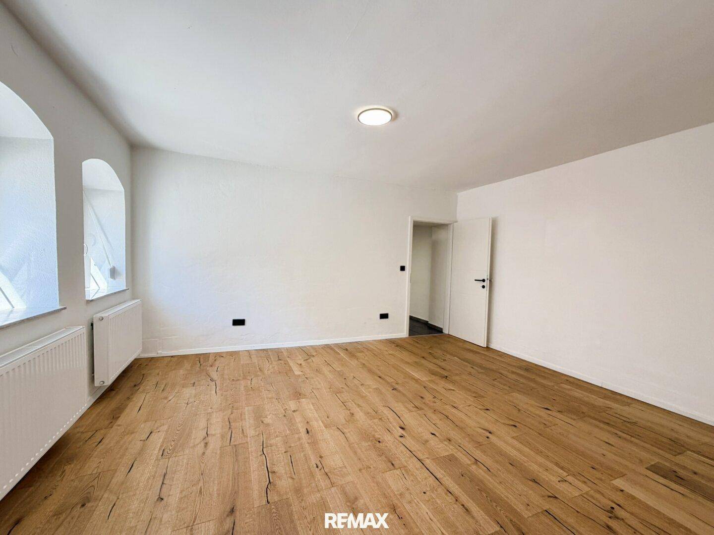 Wohnraum mit Holzboden, weißen Wänden und zwei charmanten Bogenfenstern, die viel Licht hereinlassen.