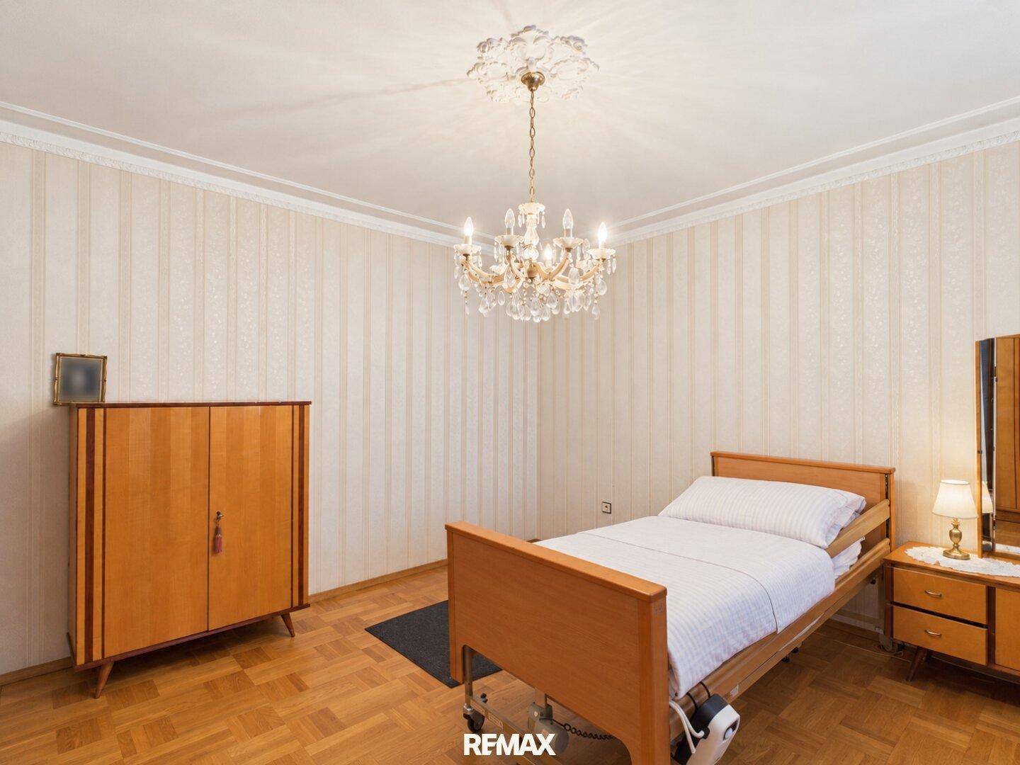 Helles Schlafzimmer mit Parkettboden, schlichter Einrichtung und einem Kronleuchter an der Decke.