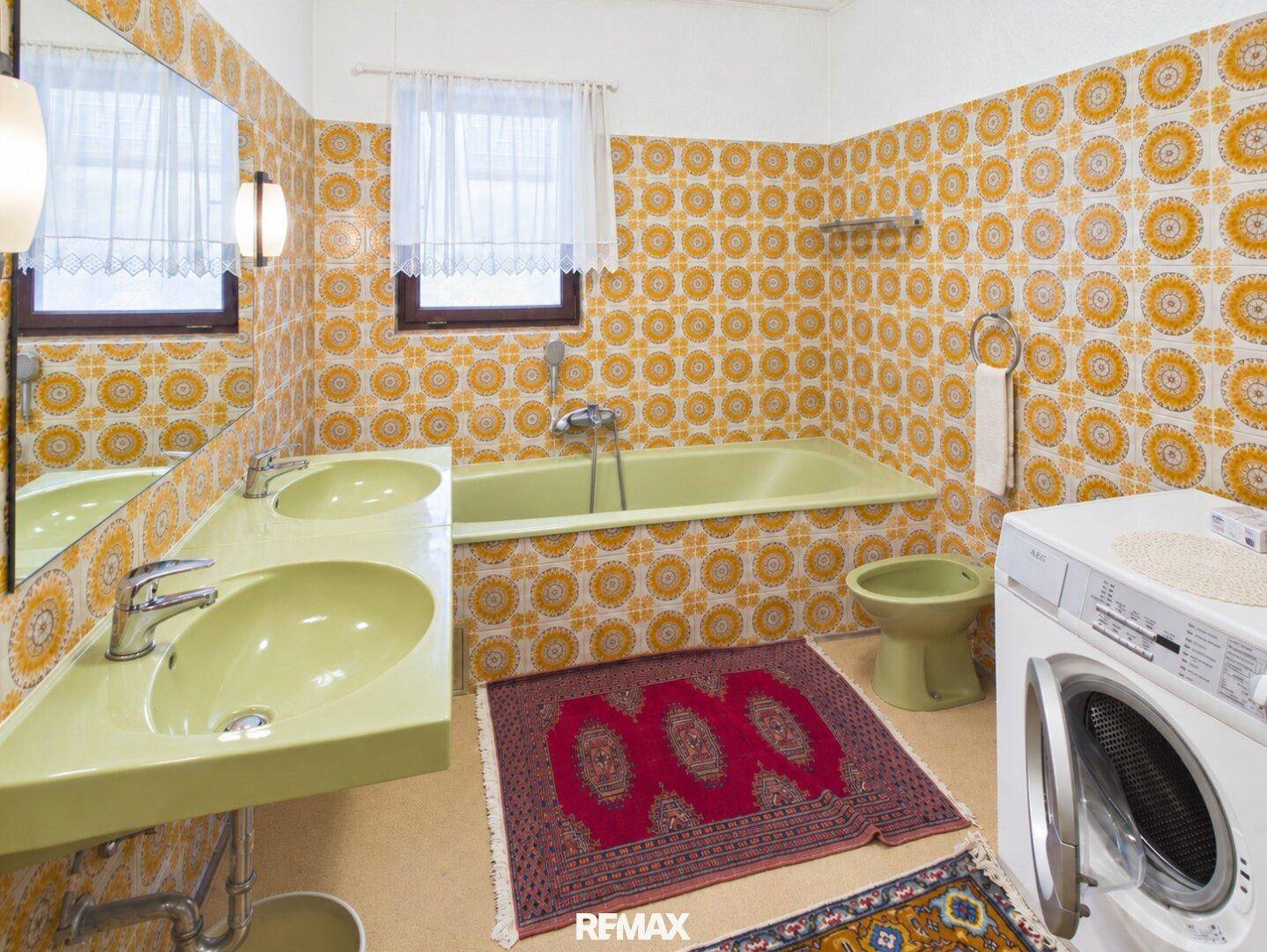 Badezimmer im Stil der 80er Jahre mit gelb-orangen Fliesen, Doppelwaschbecken und einer Badewanne.