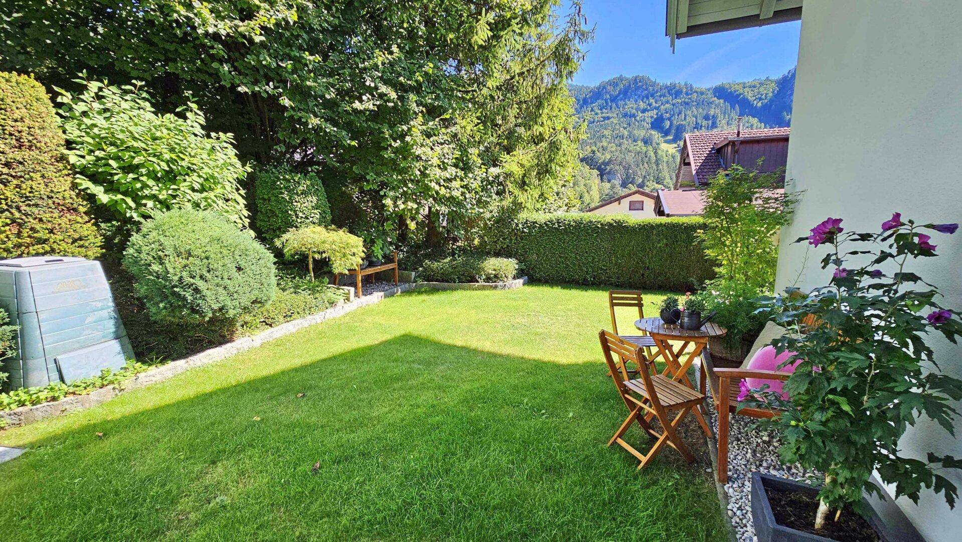 Gepflegter Garten mit Rasenfläche und Sitzgelegenheiten, umgeben von Hecken und Bäumen.