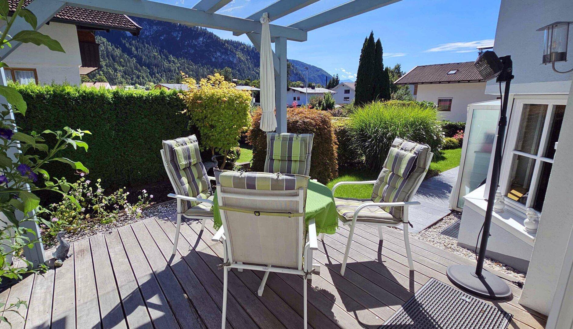 Sonniger Terrassenbereich mit Holzdeck, Pergola und Gartenmöbeln, umgeben von Grün.