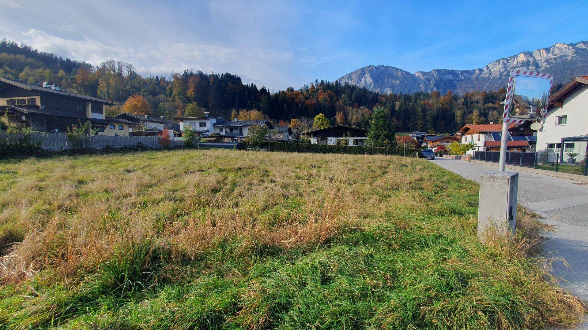 Blick über ein grünes Grundstück mit Häusern und Bergen im Hintergrund unter blauem Himmel.
