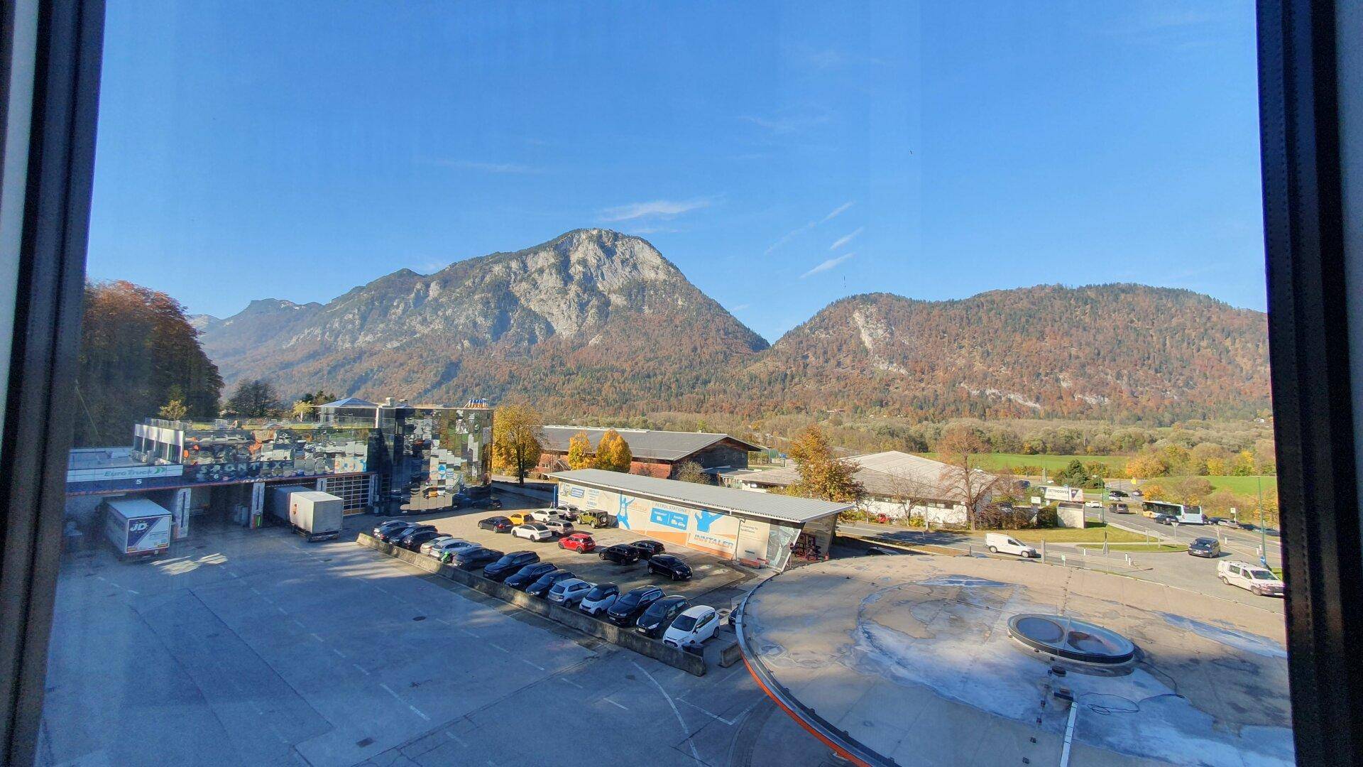 Panoramablick aus dem Fenster auf eine Berglandschaft, einen Parkplatz und umliegende Gebäude unter blauem Himmel.