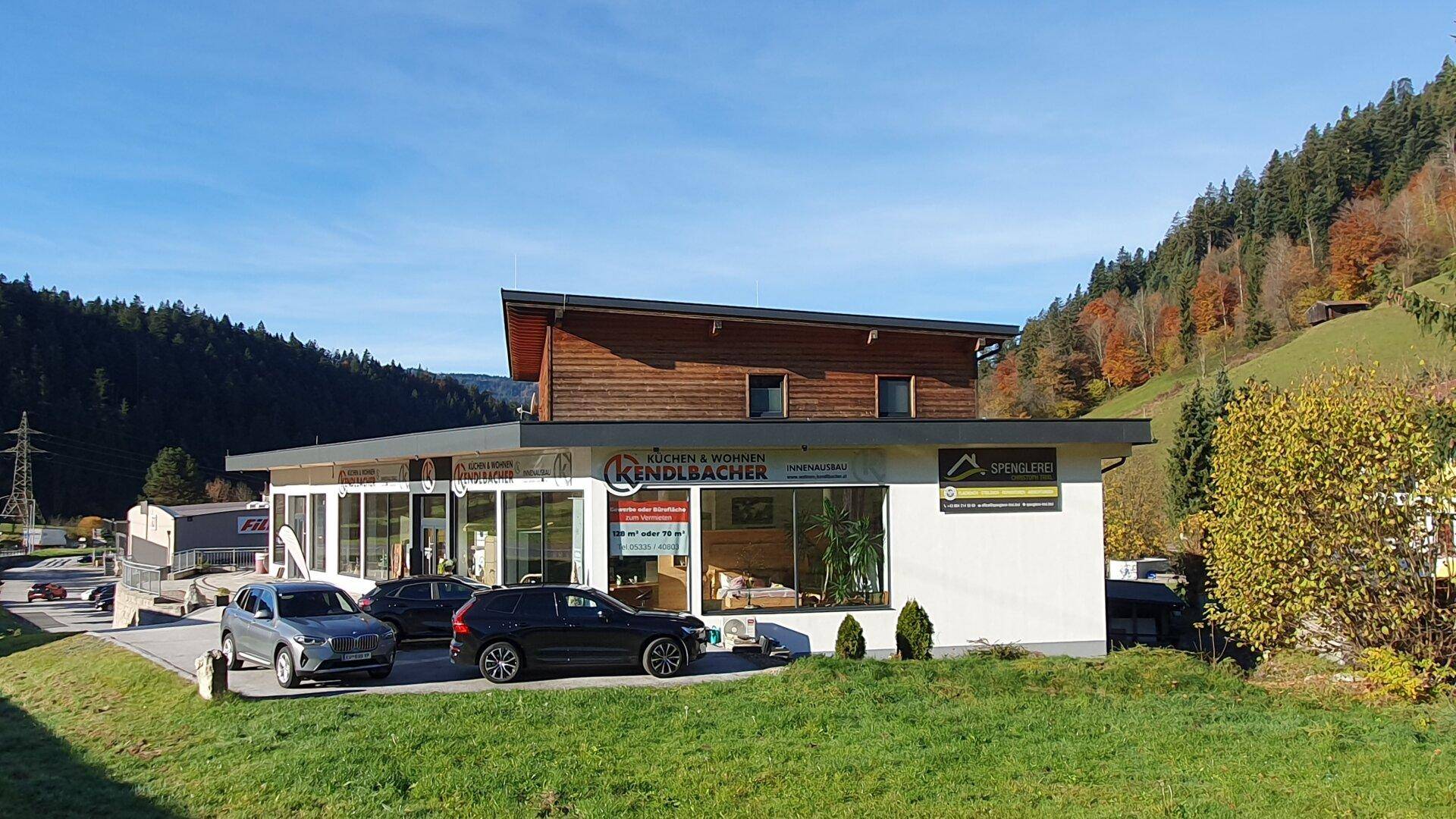 Modernes Geschäftsgebäude mit großen Fenstern und Holzfassade, umgeben von grünen Wiesen und bewaldeten Hügeln.