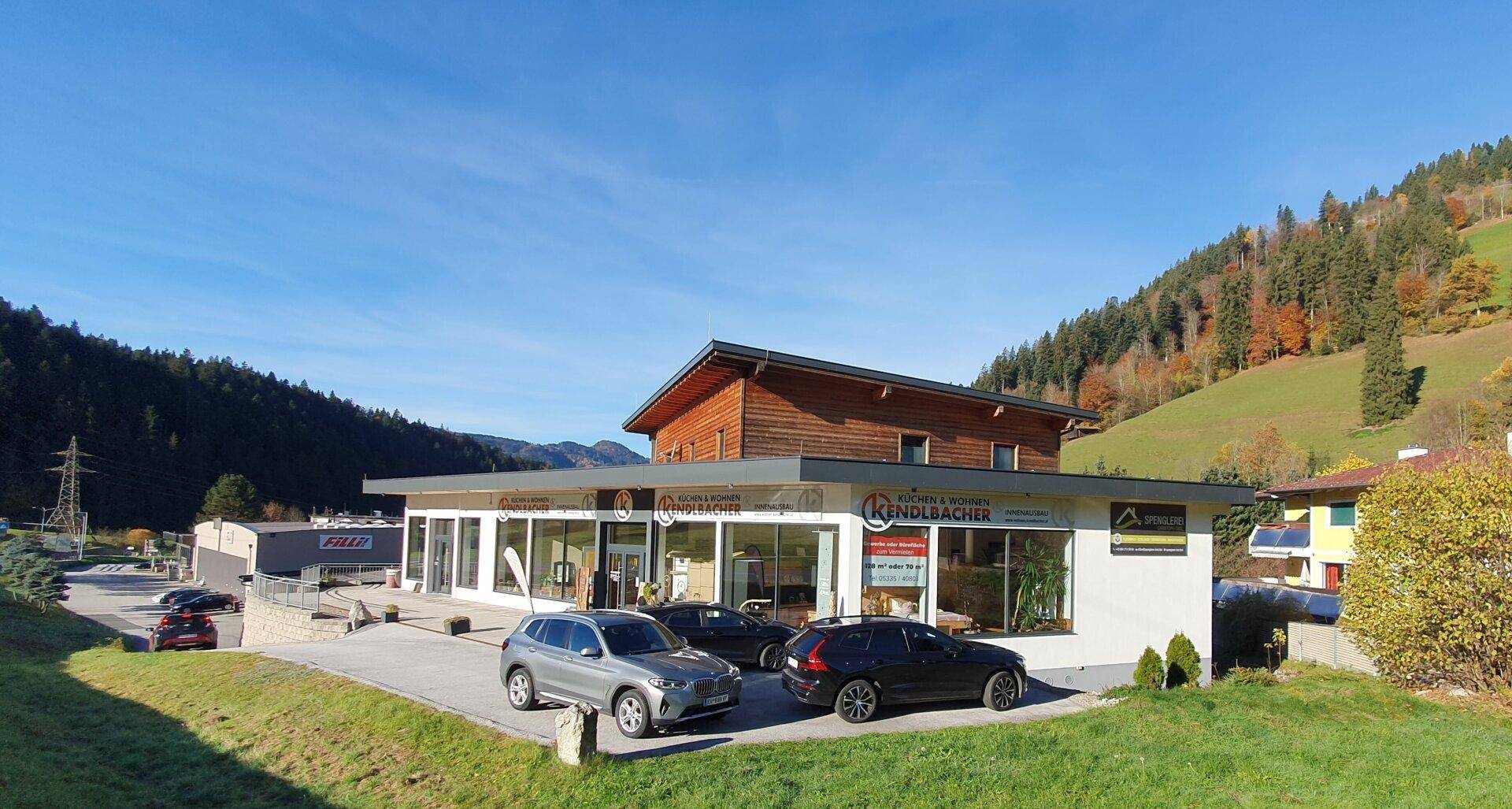 Seitenansicht des modernen Gebäudes mit Holzverkleidung und Parkplätzen, eingebettet in eine grüne Hügellandschaft.