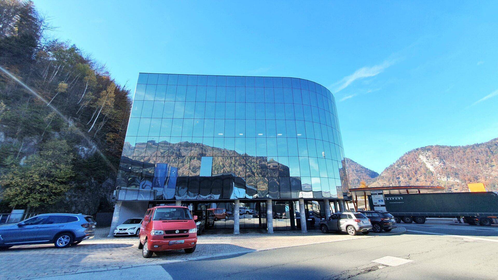 Gebäude mit geschwungener Glasfassade und reflektierenden Oberflächen, umgeben von Natur und Fahrzeugen.
