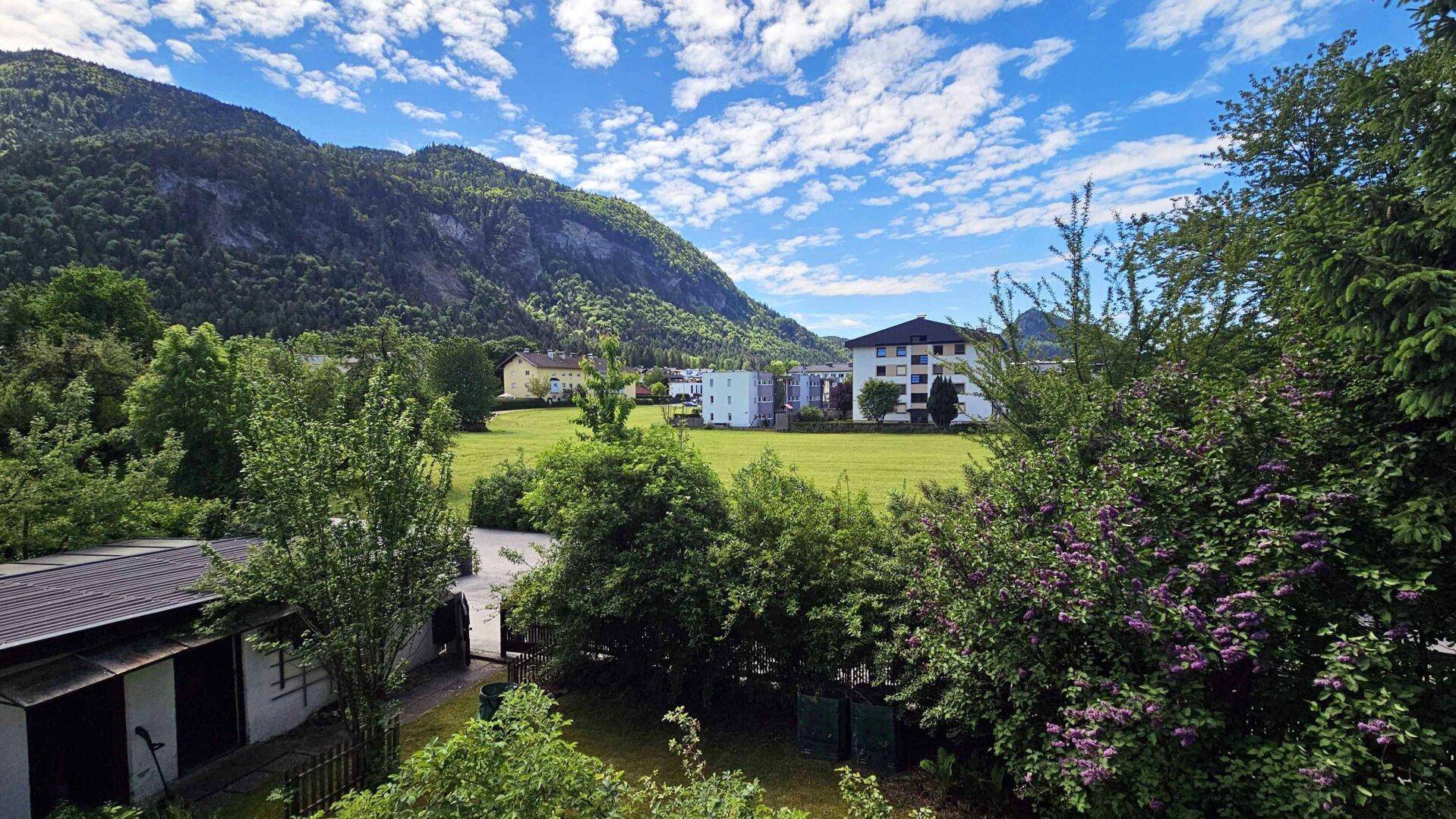 Weitläufiger Blick über grüne Felder und Berge mit einigen Gebäuden im Hintergrund unter blauem Himmel.