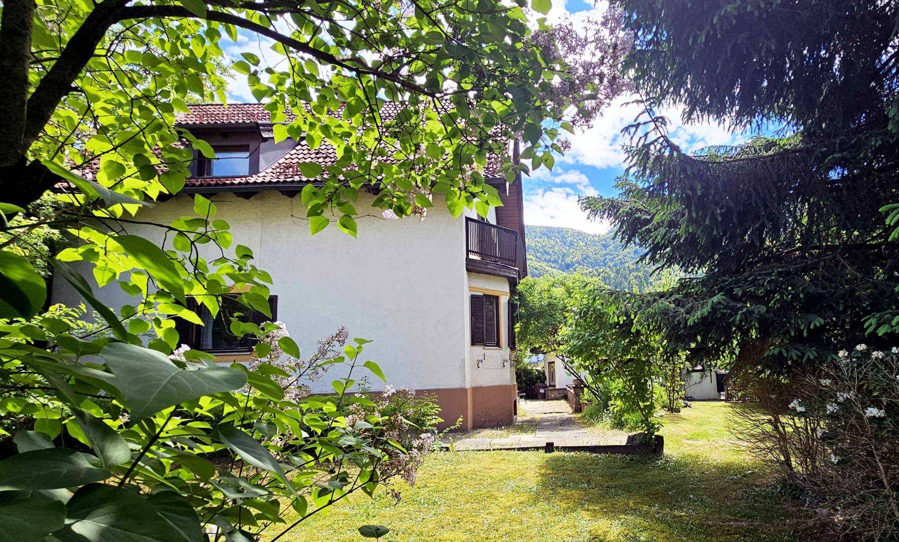 Seitenansicht des Hauses mit Balkon, umgeben von üppigem Grün und Bergblick.