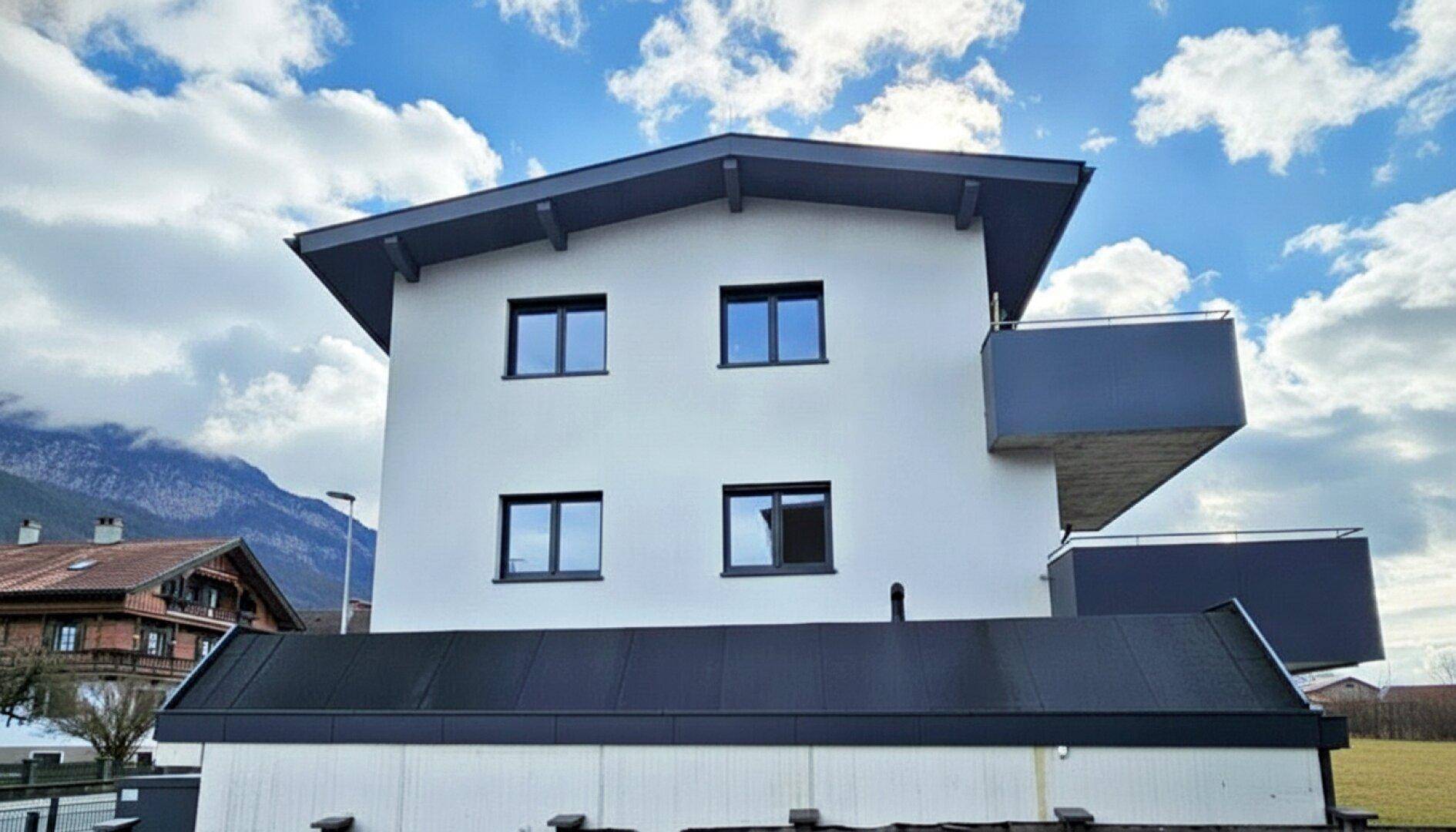 Modernes Gebäude mit schrägem Dach, Balkon und Bergblick unter bewölktem Himmel.
