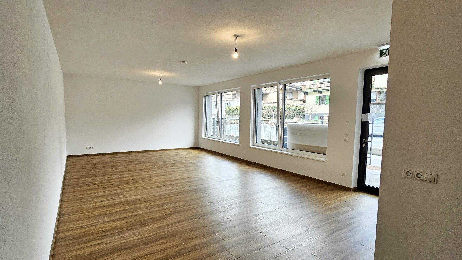 Heller, leerer Wohnraum mit großen Fenstern, Holzboden und Zugang nach draußen.