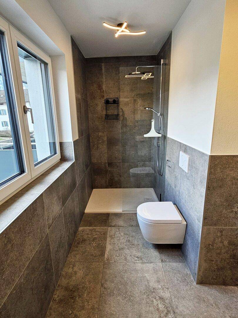 Modernes Badezimmer mit grauen Fliesen, ebenerdiger Dusche und wandhängendem WC.