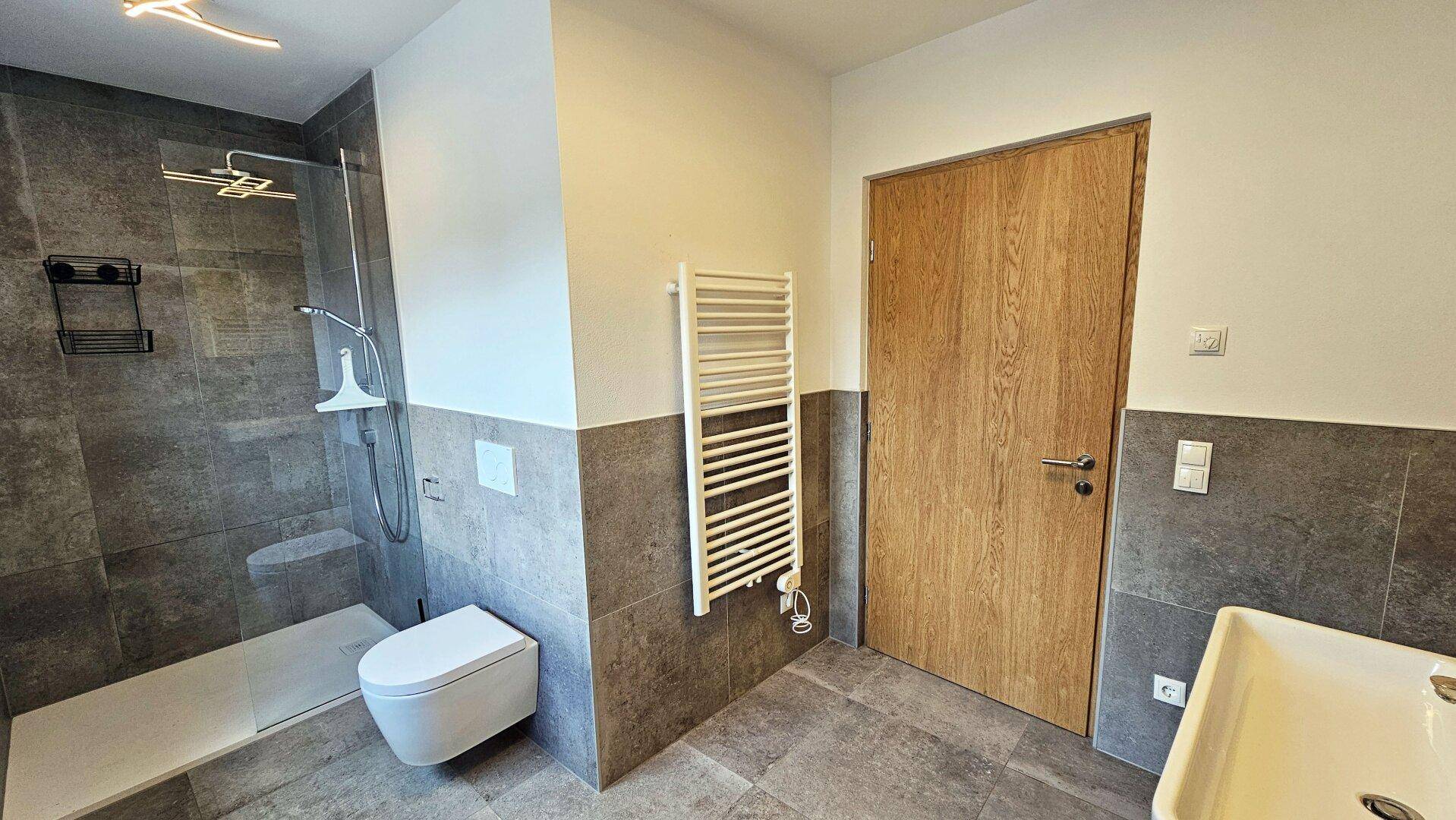 Modernes Badezimmer mit grauen Fliesen, ebenerdiger Dusche, WC und Handtuchheizkörper.