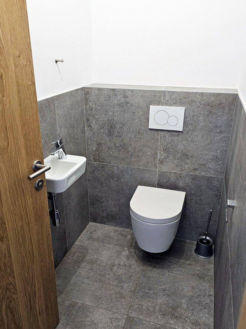 Kleines Gäste-WC mit wandhängendem WC, kleinem Waschbecken und grauen Fliesen.
