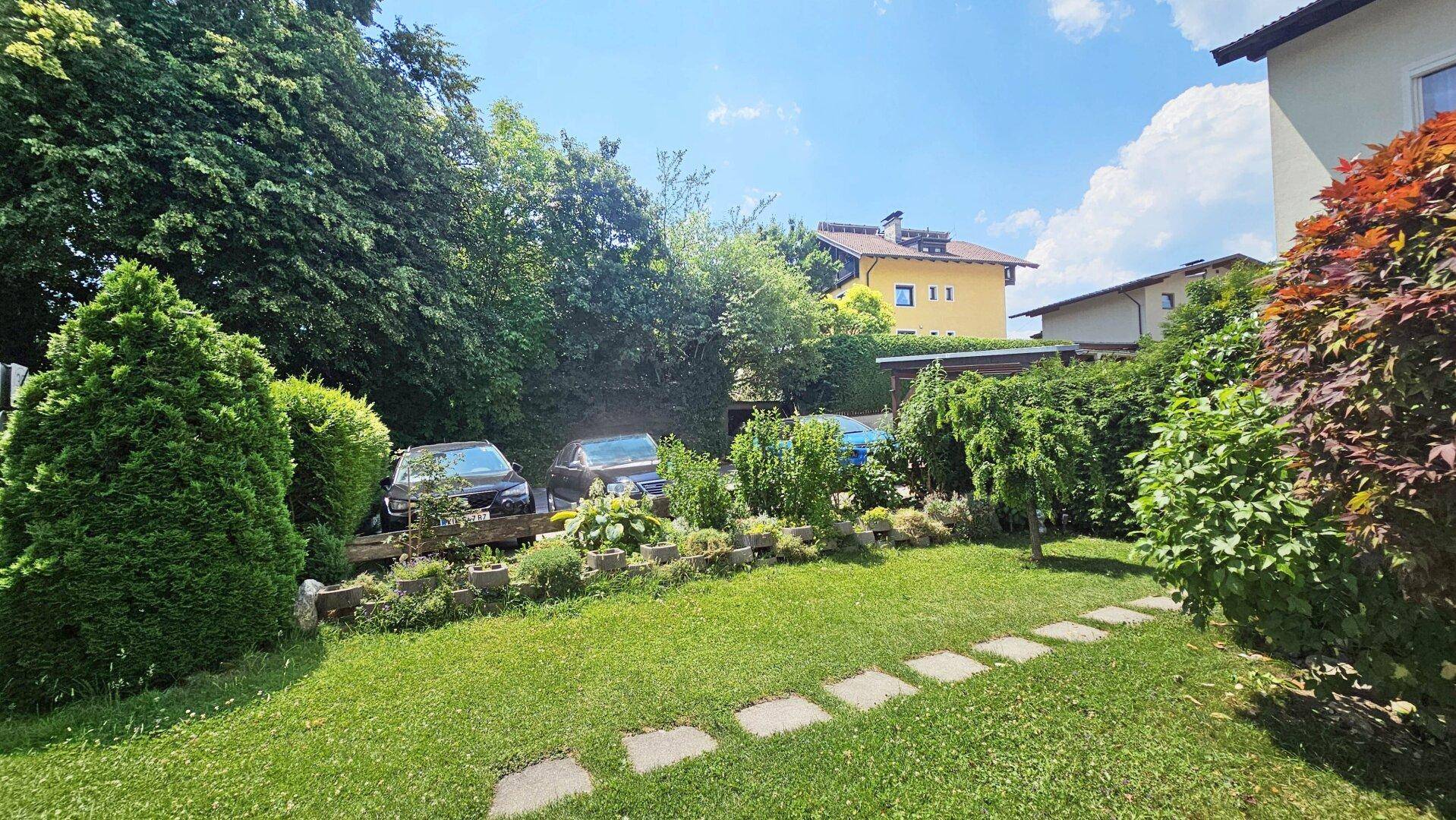 Grüner Gartenbereich mit Rasen, Bäumen und Stellplätzen im Hintergrund.