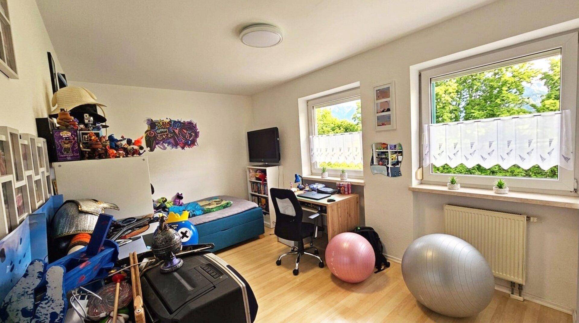 Freundliches Kinderzimmer mit Schreibtisch, Bett und zwei Fenstern mit Blick ins Grüne.