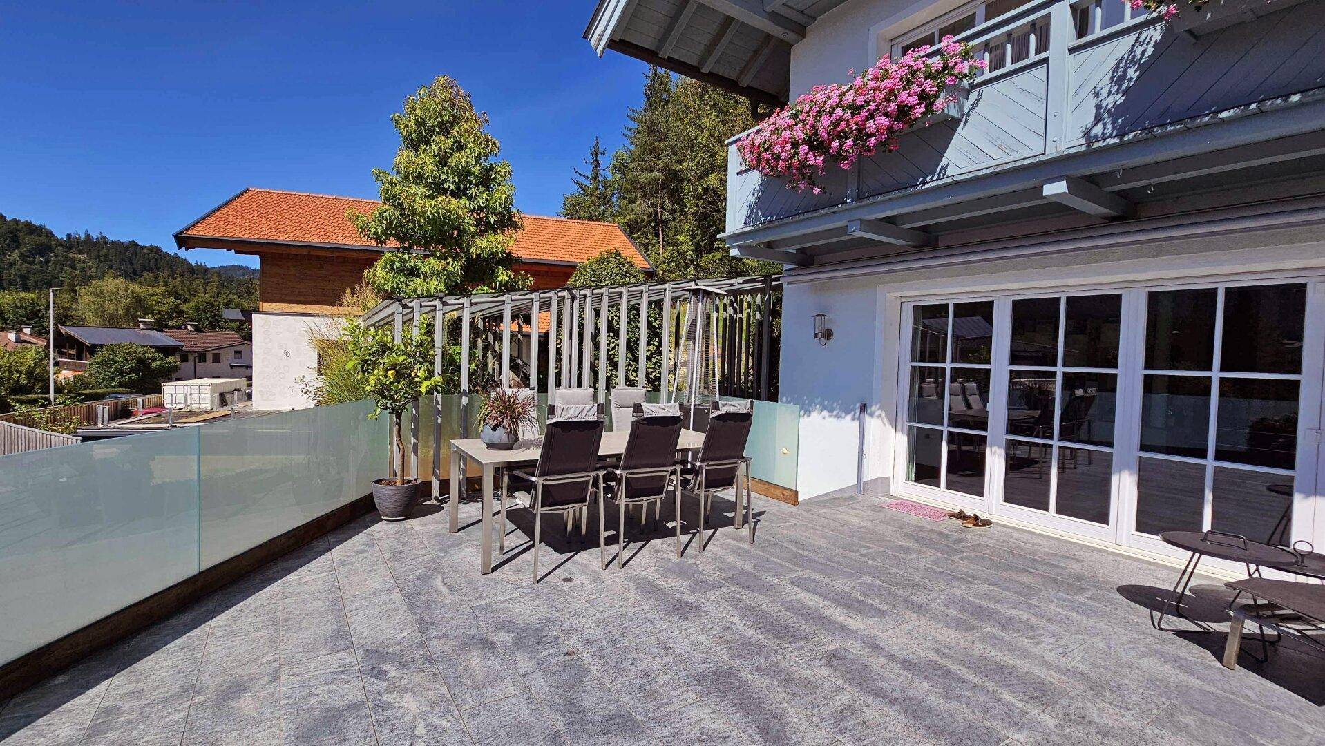 Große, sonnige Terrasse mit Essbereich und Glasgeländer, umgeben von Häusern und Bergen.