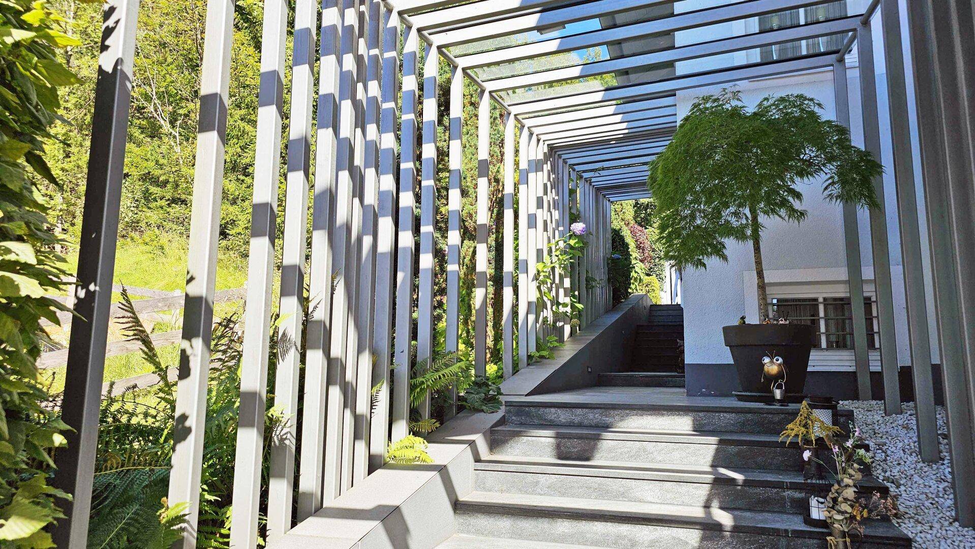 Moderner Eingangsbereich mit Pergola und Treppe, umgeben von Pflanzen und Natur.
