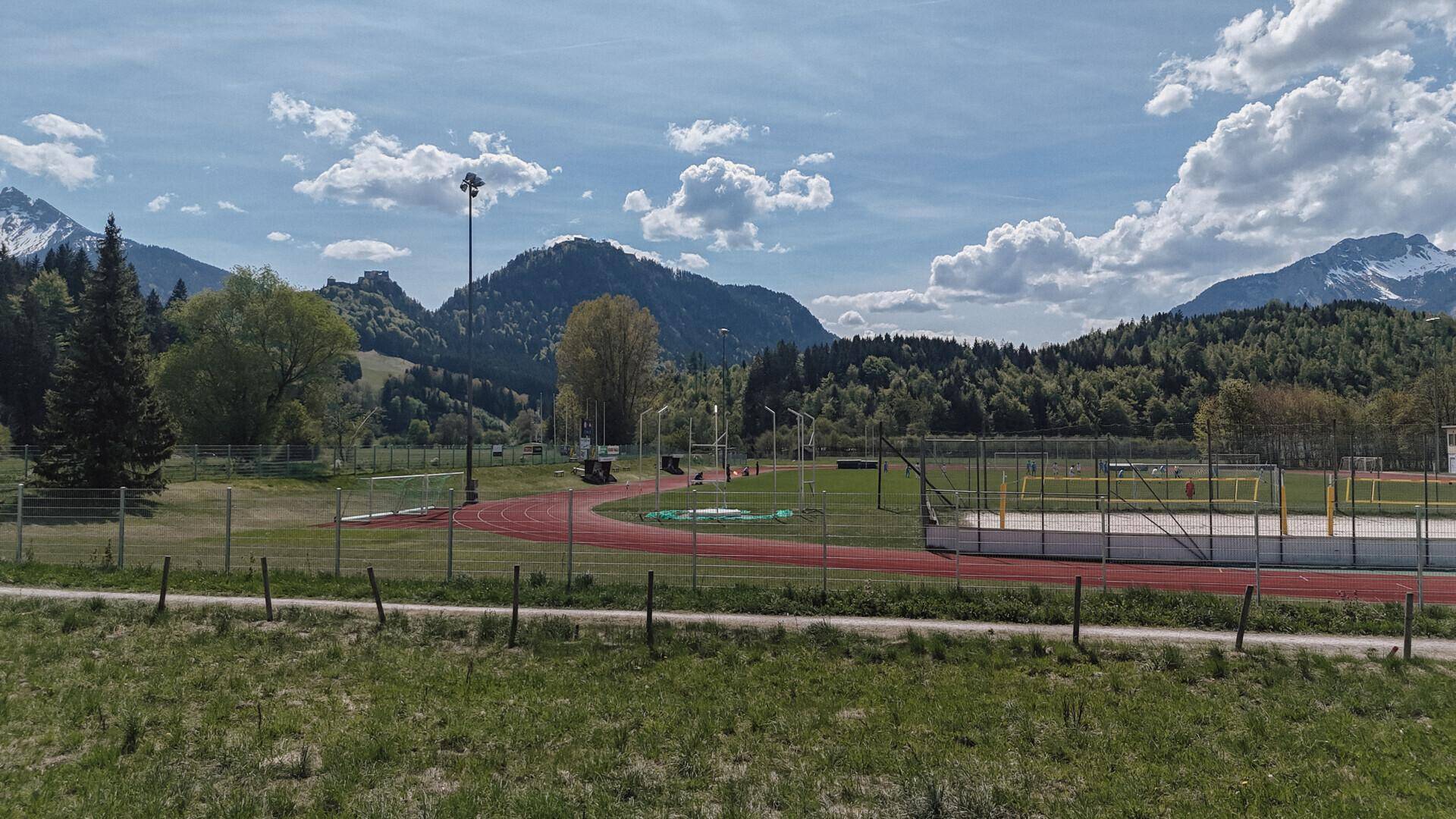 Sportanlage mit Laufbahn und Spielfeld, umgeben von grünen Hügeln und Bergen unter einem bewölkten Himmel.