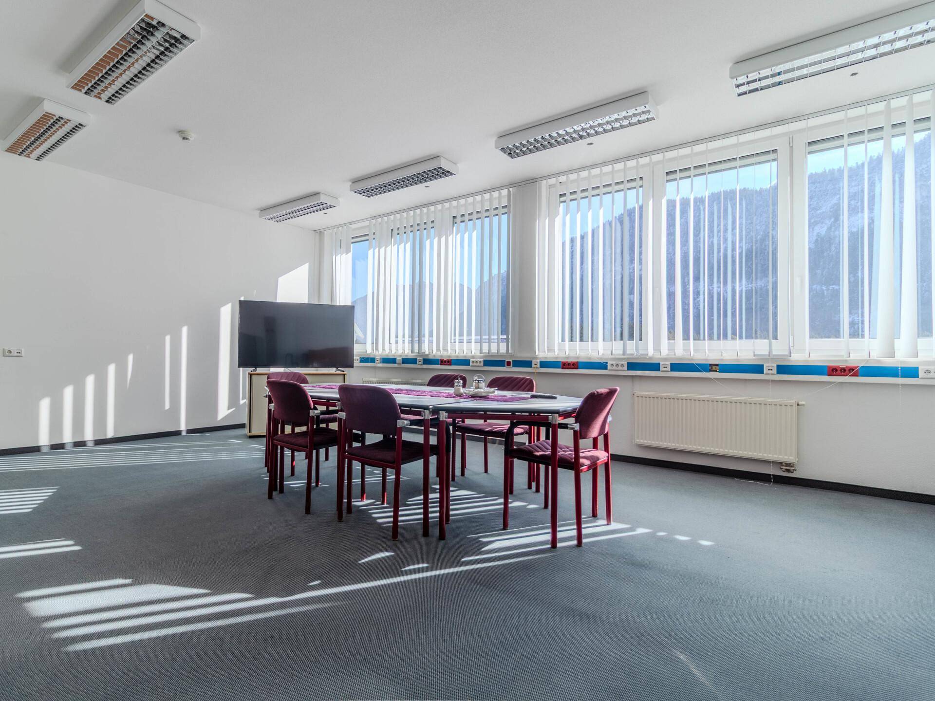 Heller Konferenzraum mit großem Tisch, Stühlen, Bildschirm und vielen Fenstern mit Jalousien.