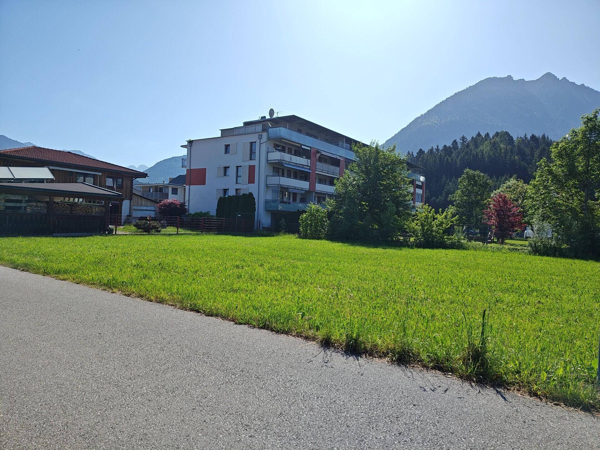 Grüne Wiese mit einem Mehrfamilienhaus und bewaldeten Bergen unter strahlend blauem Himmel.