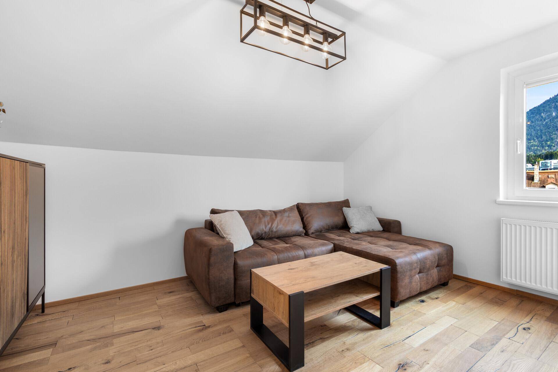 Helles Wohnzimmer mit braunem Ecksofa, Holzboden und Fenster mit Bergblick.