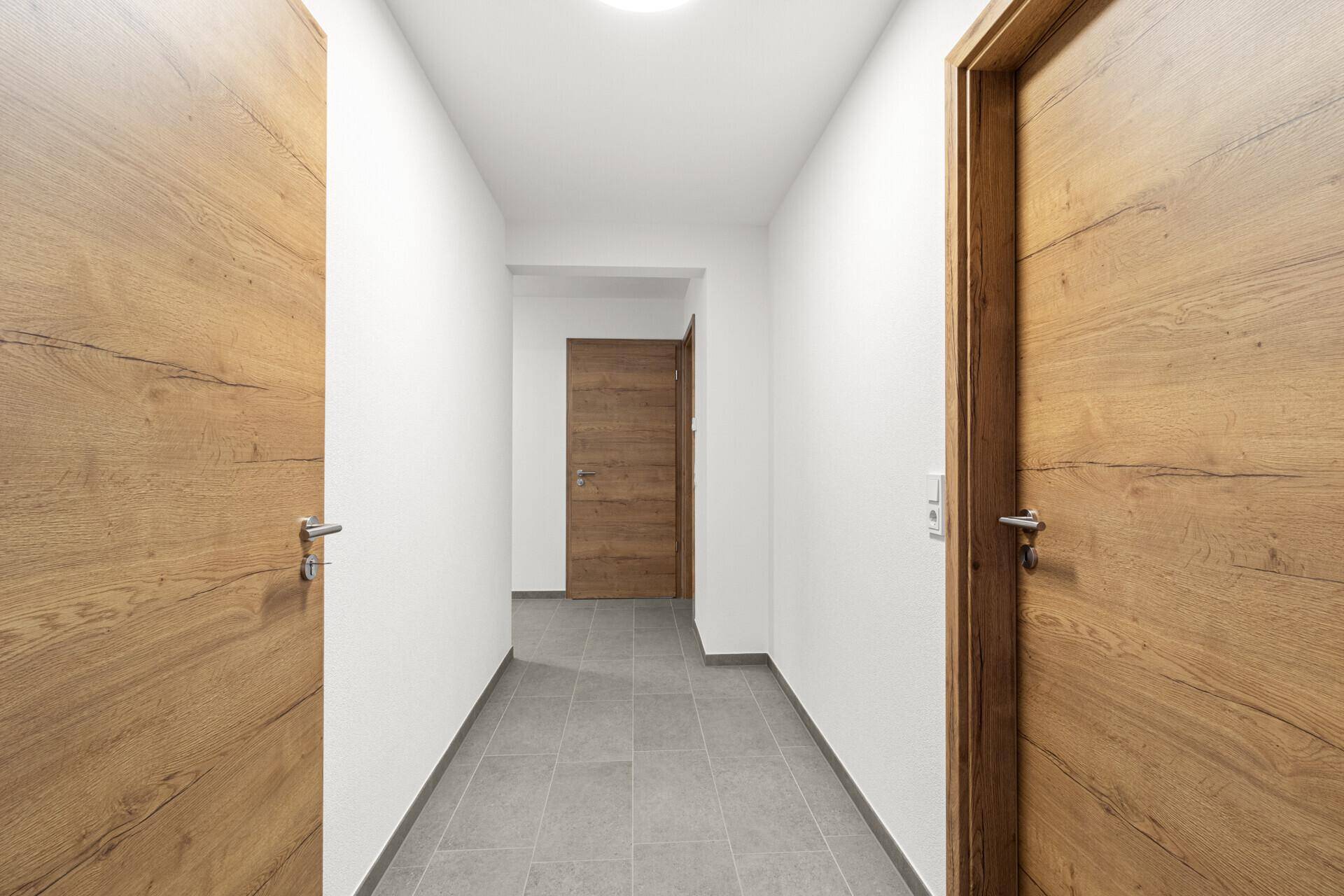 Schmaler Flur mit Fliesenboden und Holztüren, der verschiedene Räume miteinander verbindet.
