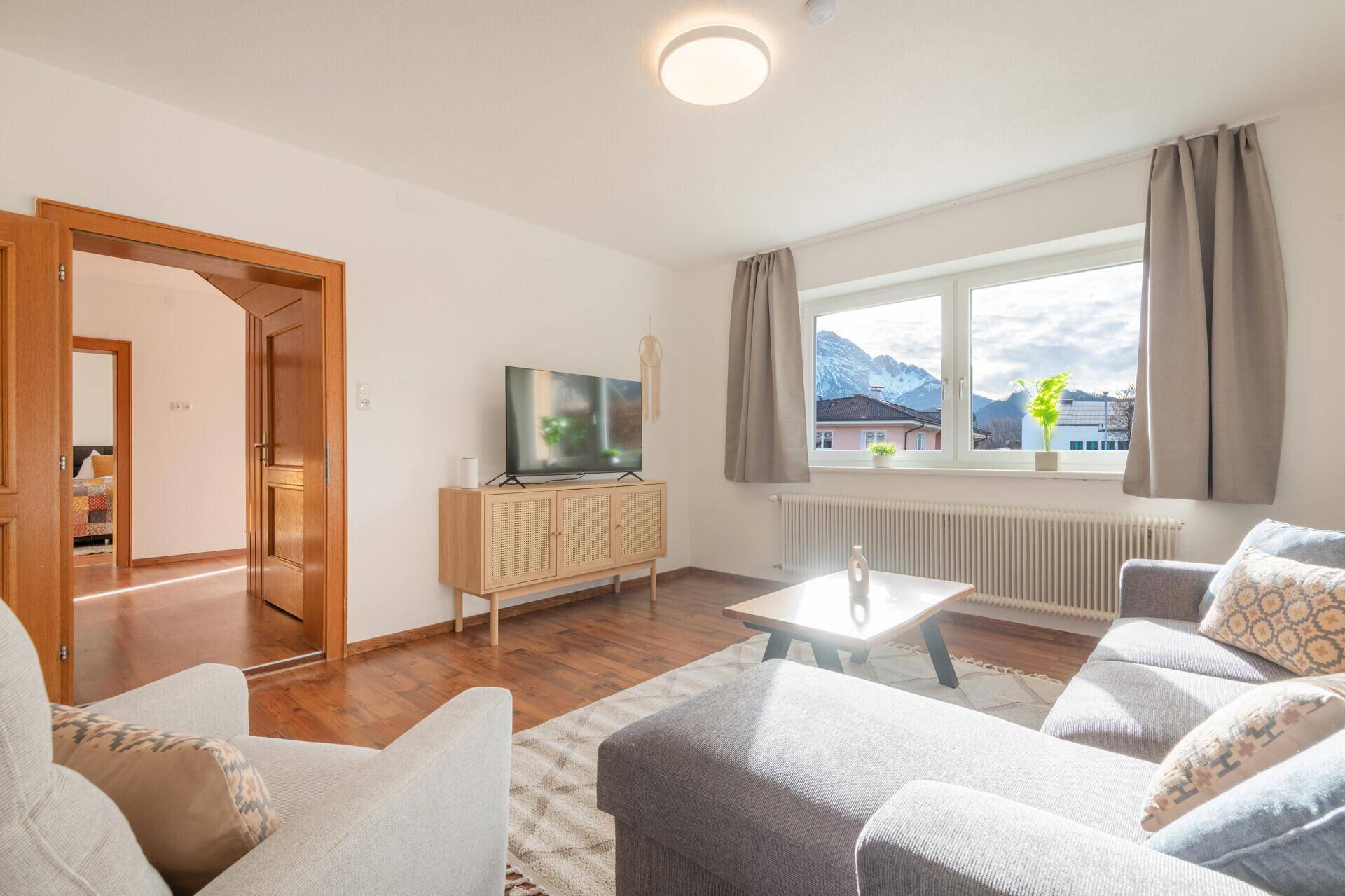Helles Wohnzimmer mit Holzboden, Fernseher und Zugang zu weiteren Räumen.