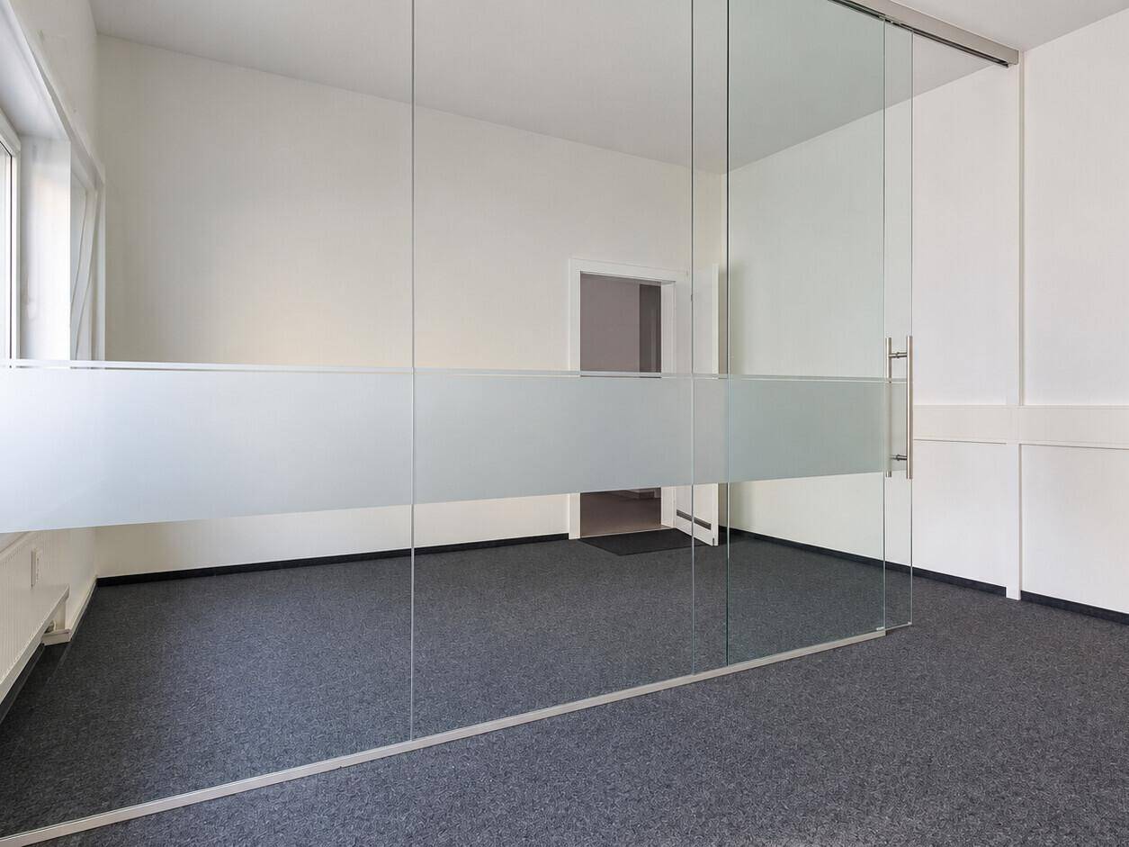 Moderner Büroraum mit Glasabtrennung, Teppichboden und weiß gestrichenen Wänden.