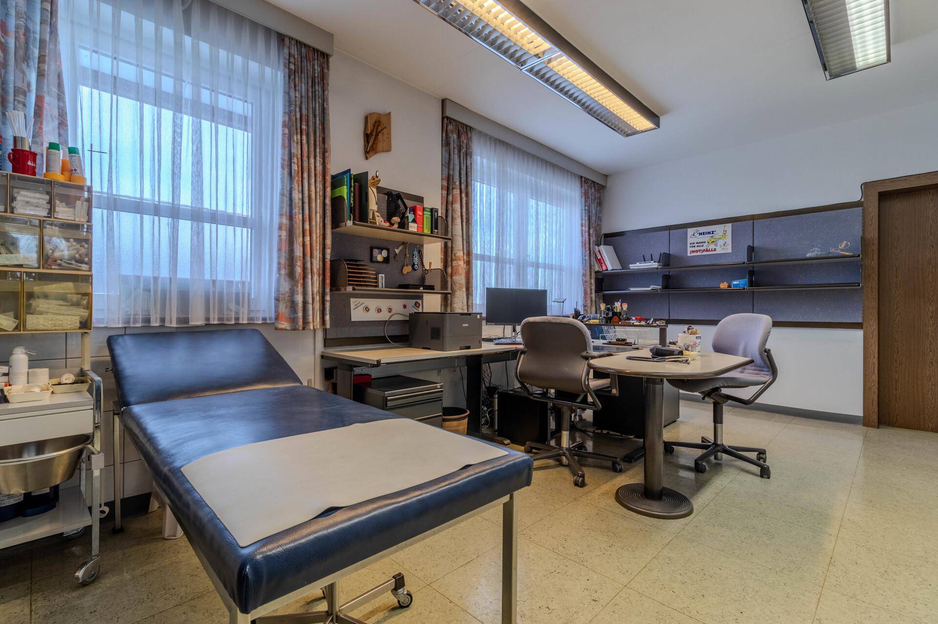 Geräumiges Behandlungszimmer mit Untersuchungsliege, Schreibtisch und Regalen, gut beleuchtet durch große Fenster.