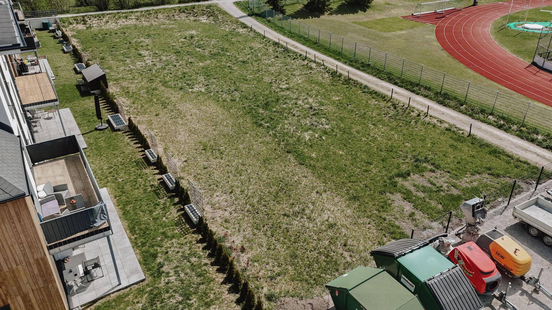 Weitläufiges grünes Grundstück neben modernen Wohngebäuden mit Balkonen und einer Sportanlage im Hintergrund.