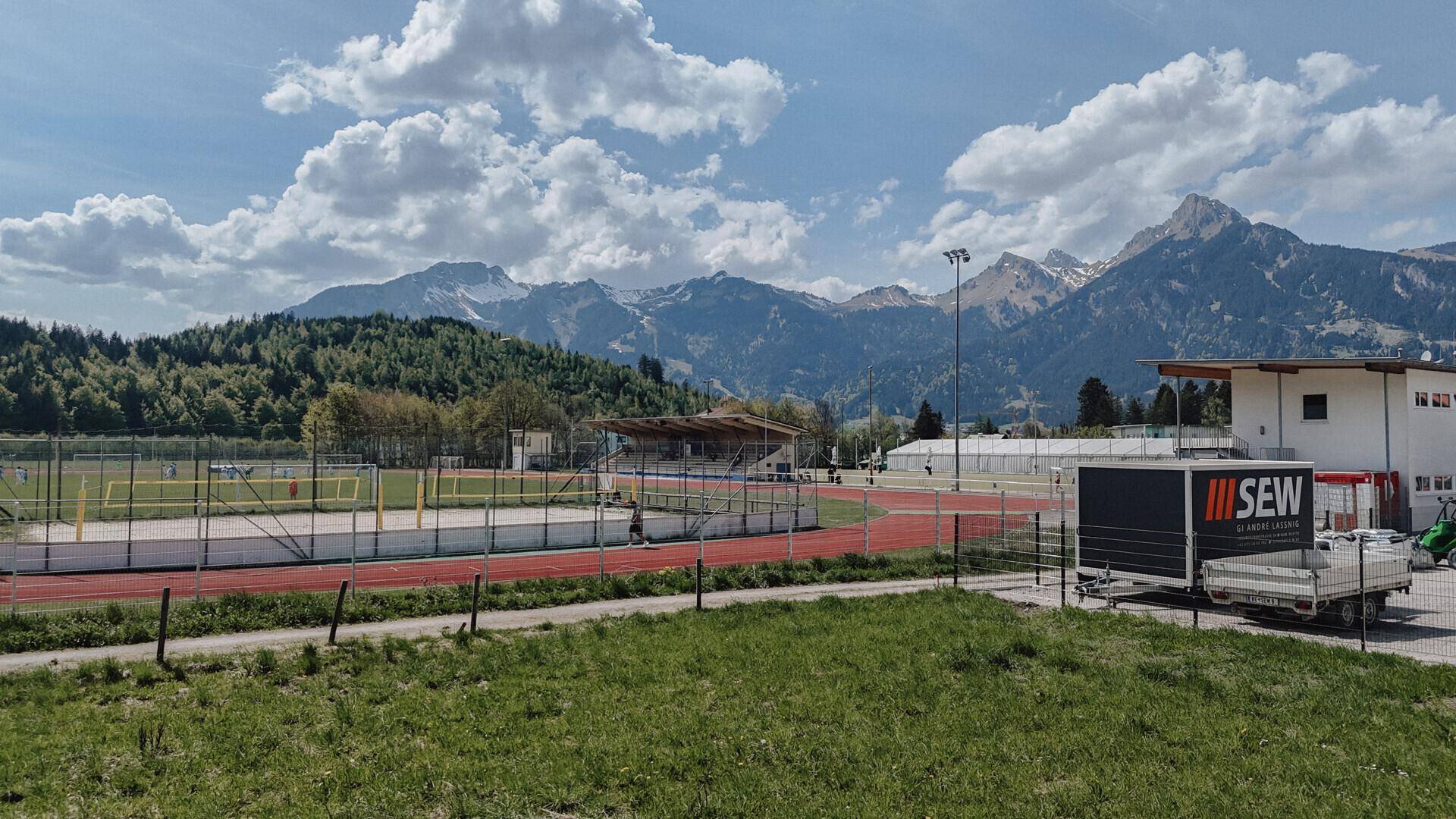 Sportanlage mit Laufbahn und Spielfeld, umgeben von einer beeindruckenden Bergkulisse und einem Gebäude rechts.