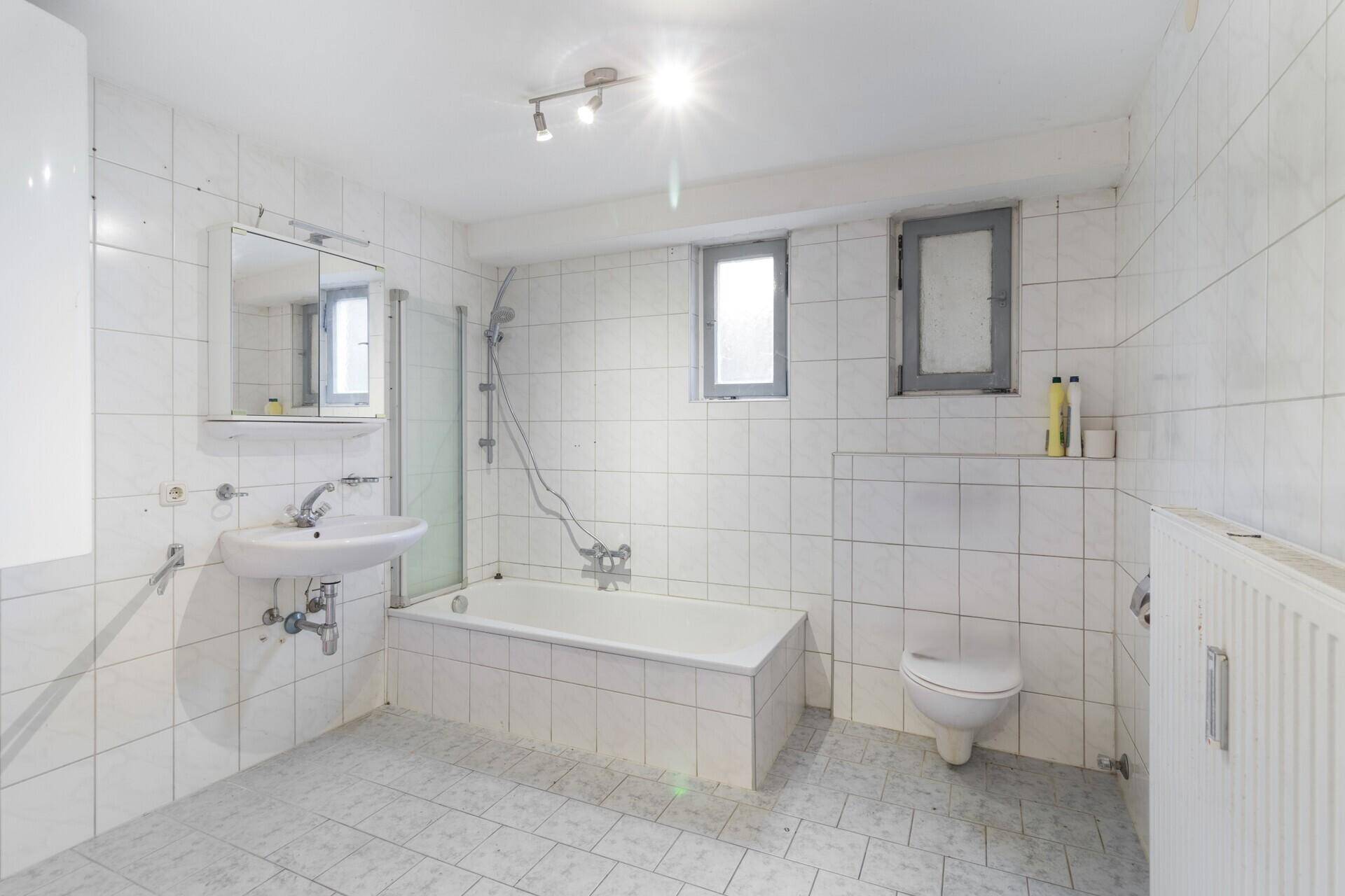 Gefliestes Badezimmer mit Badewanne, Waschbecken, Toilette und zwei kleinen Fenstern.