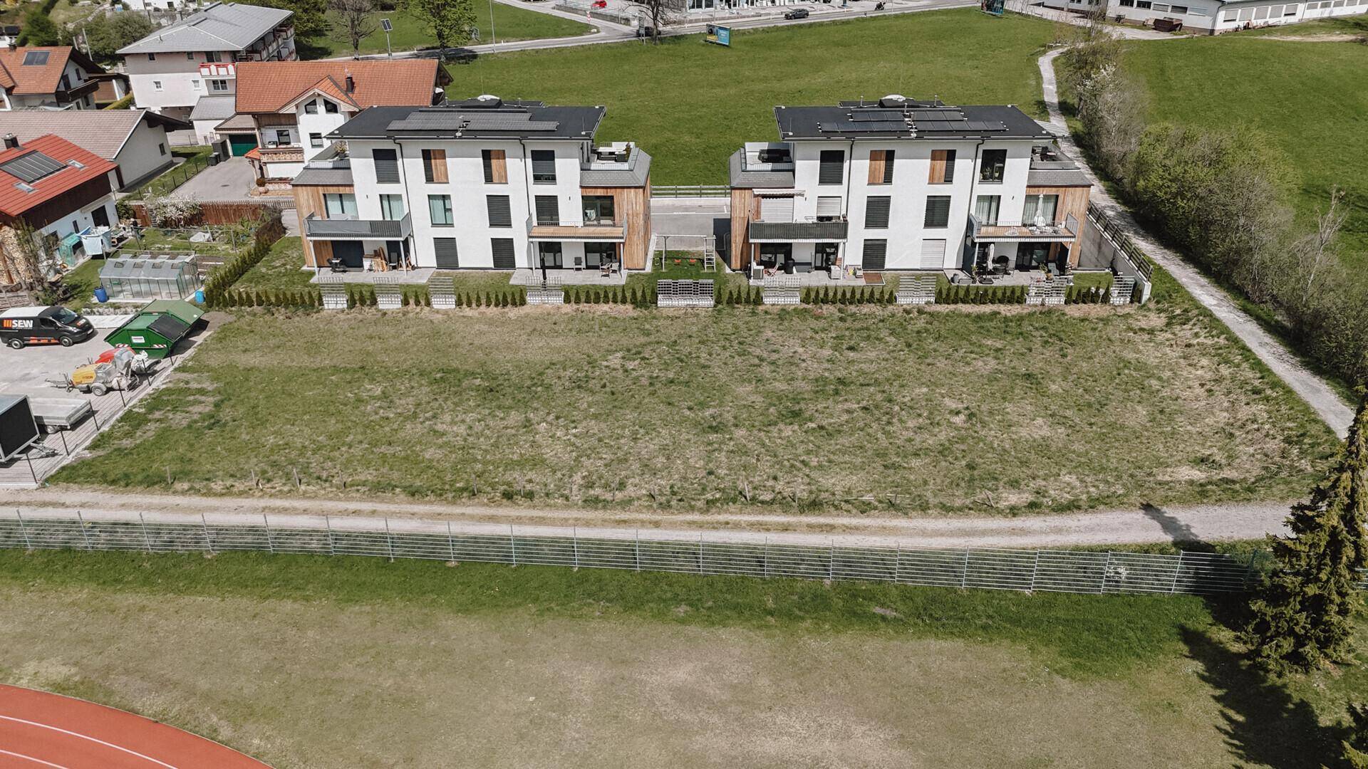 Zwei moderne Mehrfamilienhäuser mit Balkonen und Terrassen grenzen an ein großes, unbebautes Wiesengrundstück.