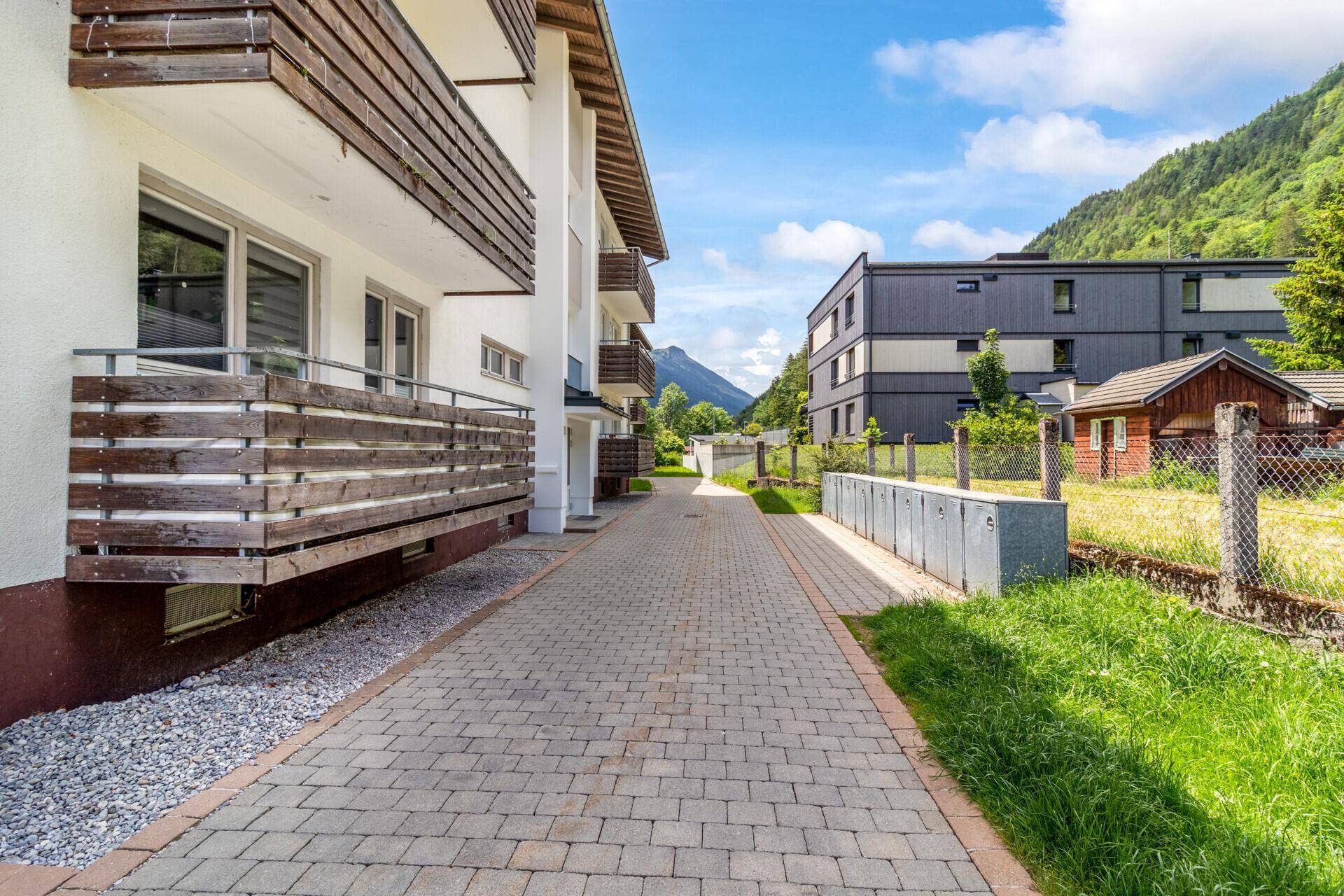 Gebäudefassade mit Balkonen und gepflastertem Weg, umgeben von grüner Landschaft.