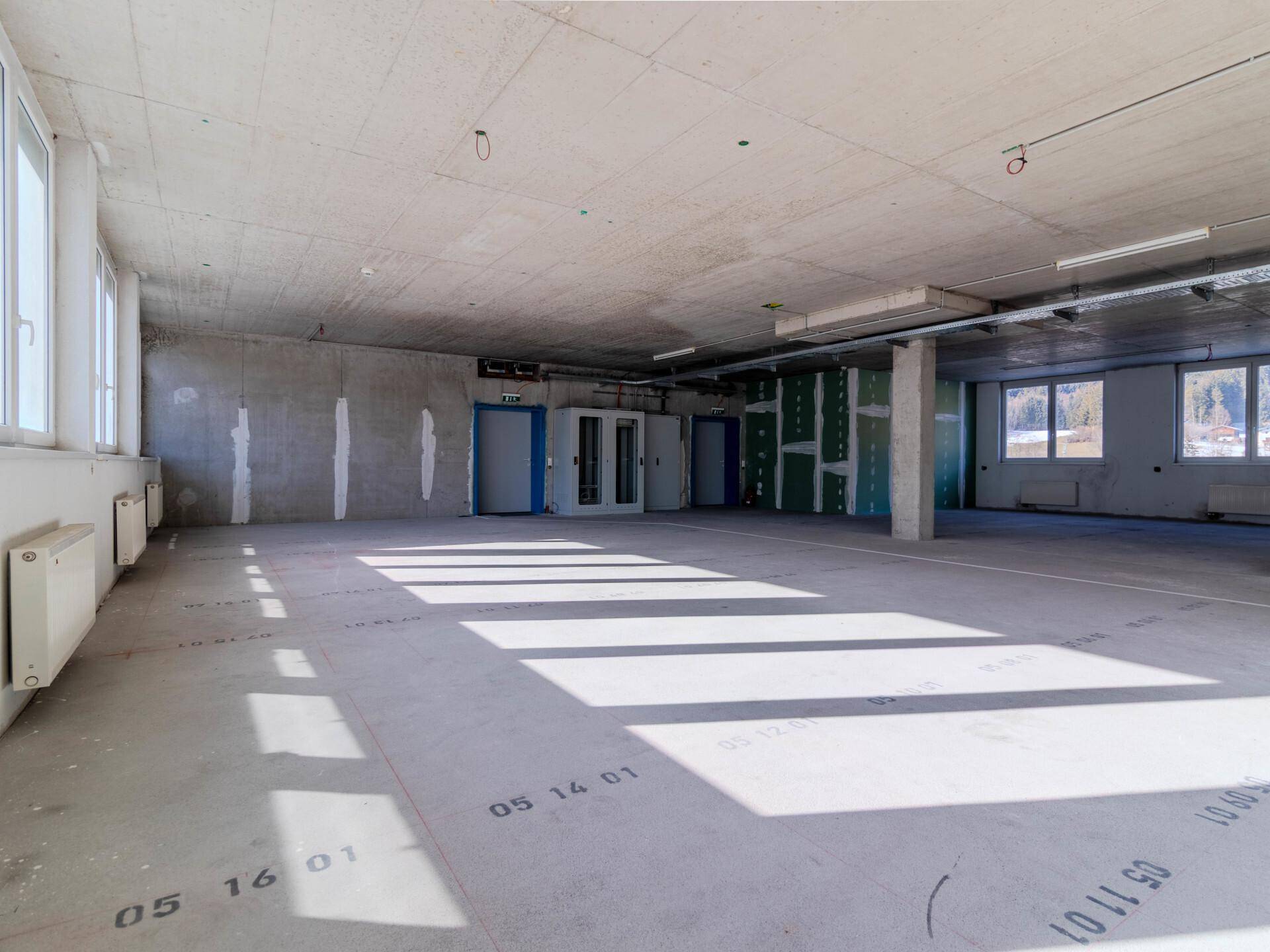 Weitläufiger, unmöblierter Bürobereich mit Betonoberflächen und natürlichem Lichteinfall durch die Fenster.