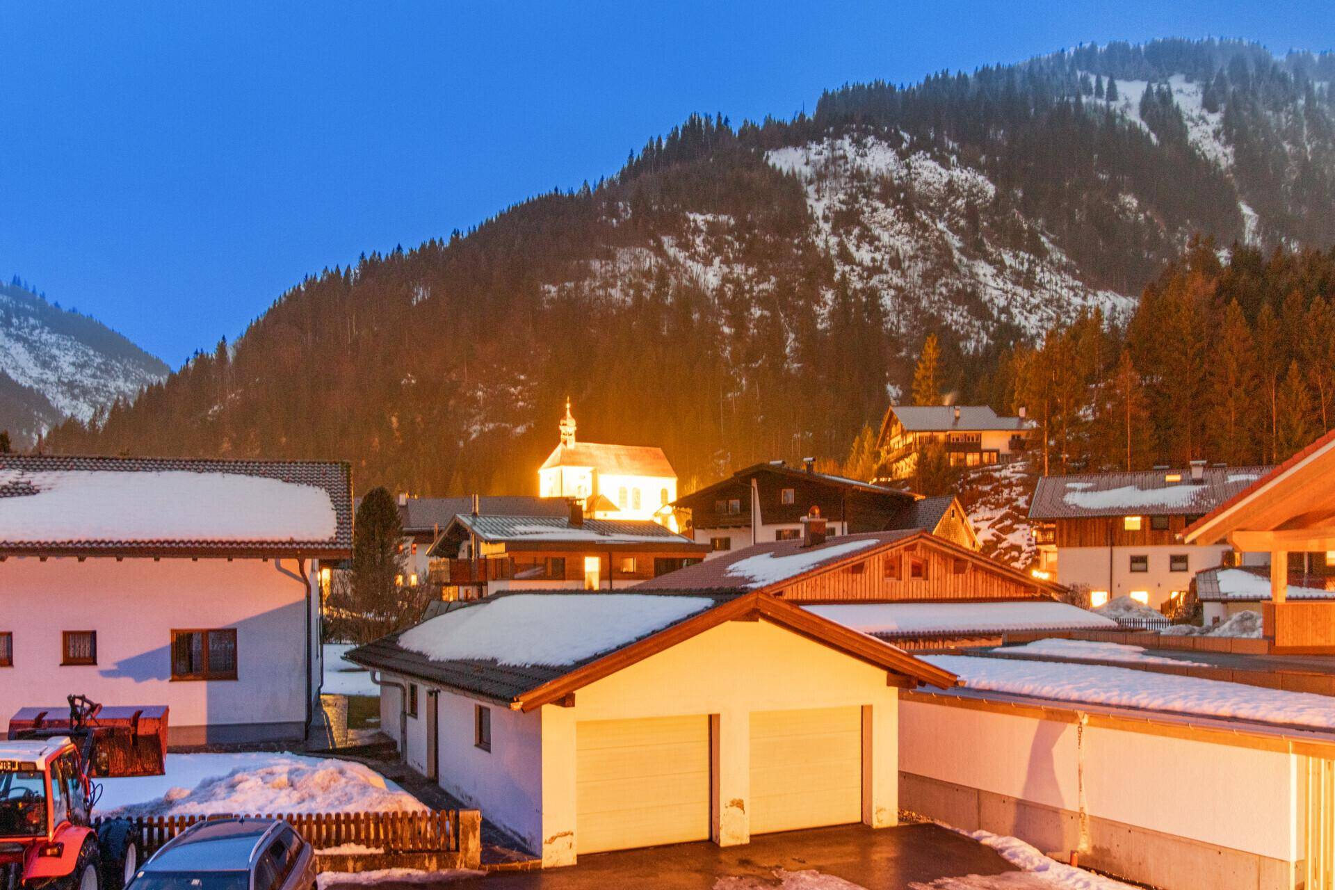 Winterliche Abendansicht eines Dorfes mit beleuchteten Häusern und schneebedeckten Bergen im Hintergrund.