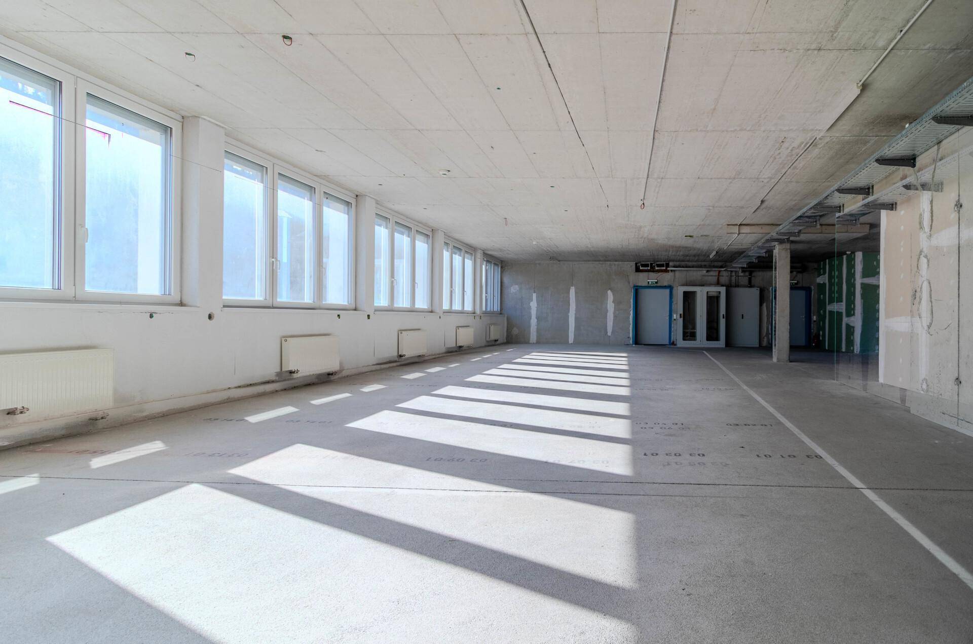 Großer, leerer Büroraum im Rohbau mit vielen Fenstern und Betonboden, bereit für den Ausbau.