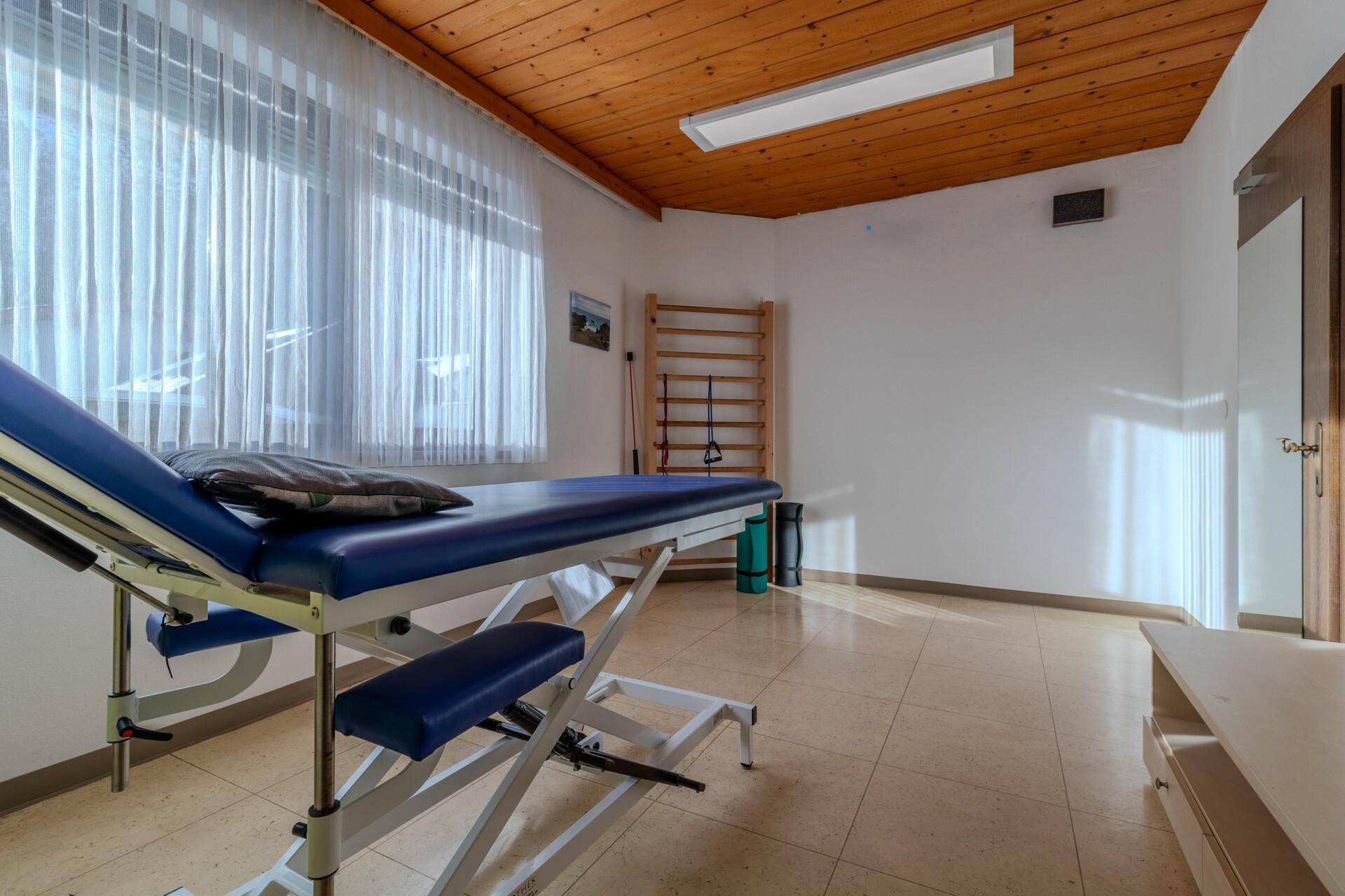 Geräumiger Therapieraum mit viel Tageslicht, Holzbalkendecke und einer modernen Behandlungsliege.