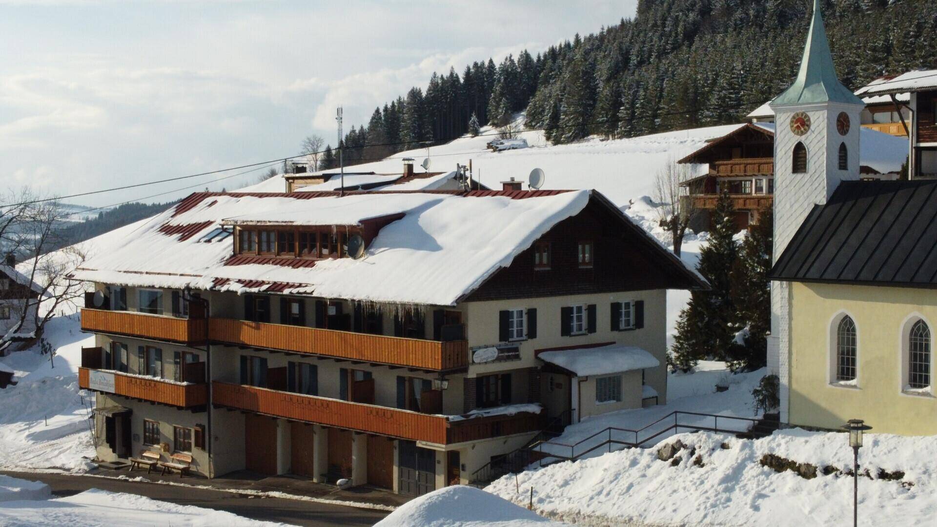 Gebäude in einem verschneiten Bergdorf mit Kirche und umliegenden Wäldern unter blauem Himmel.