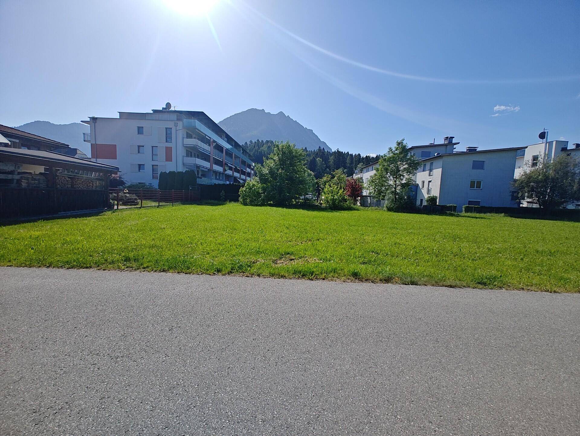 Weitläufige grüne Wiese vor mehreren Mehrfamilienhäusern mit Bergen im Hintergrund an einem sonnigen Tag.