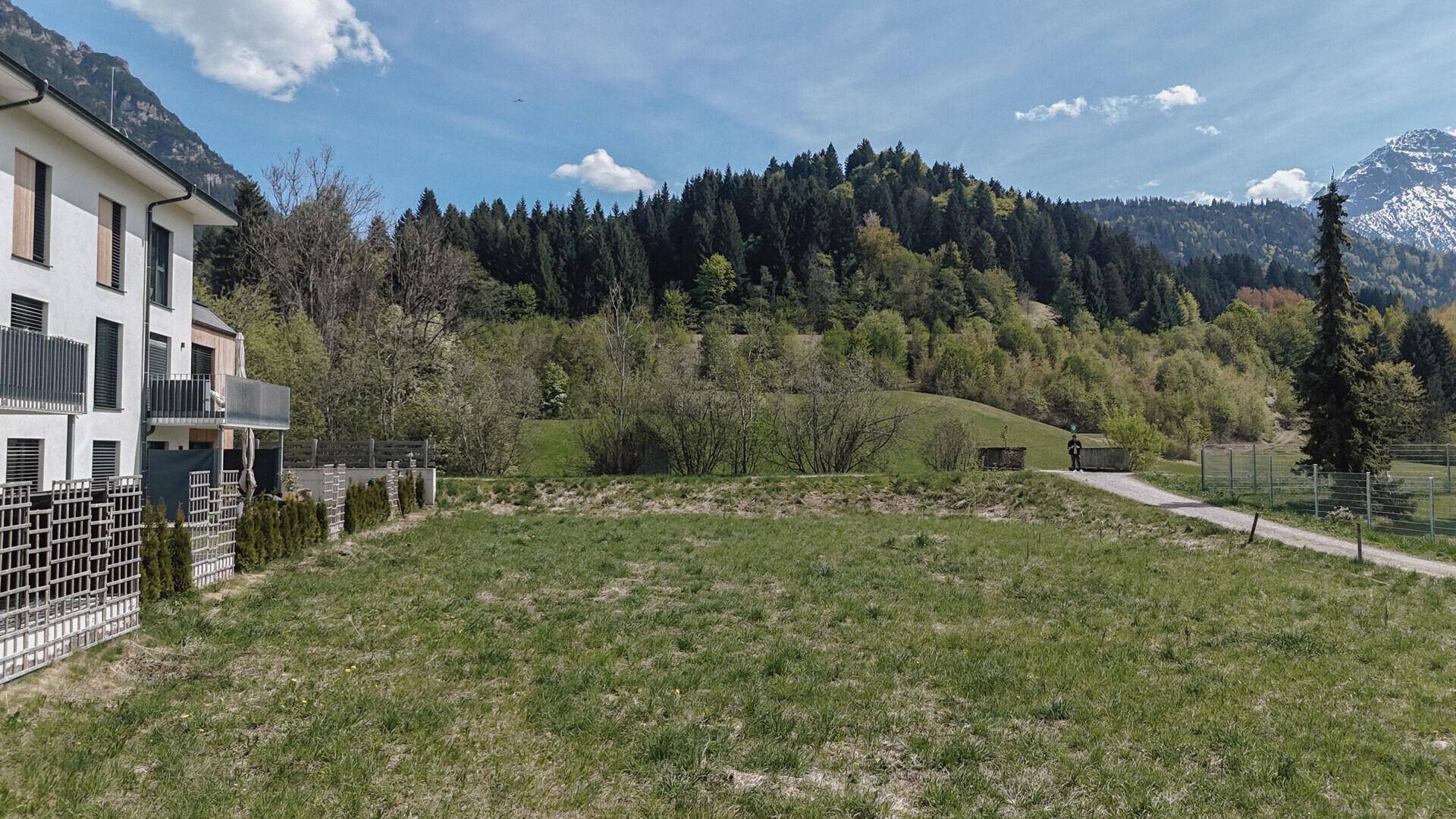 Grünes Grundstück mit modernen Gebäuden links, einem Weg und einer bewaldeten Hügellandschaft mit Bergen im Hintergrund.
