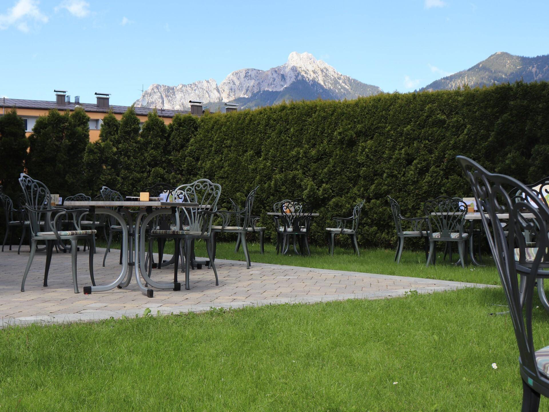 Eine sonnige Außenterrasse mit Tischen und Stühlen, umgeben von einer hohen Hecke und mit Bergblick.