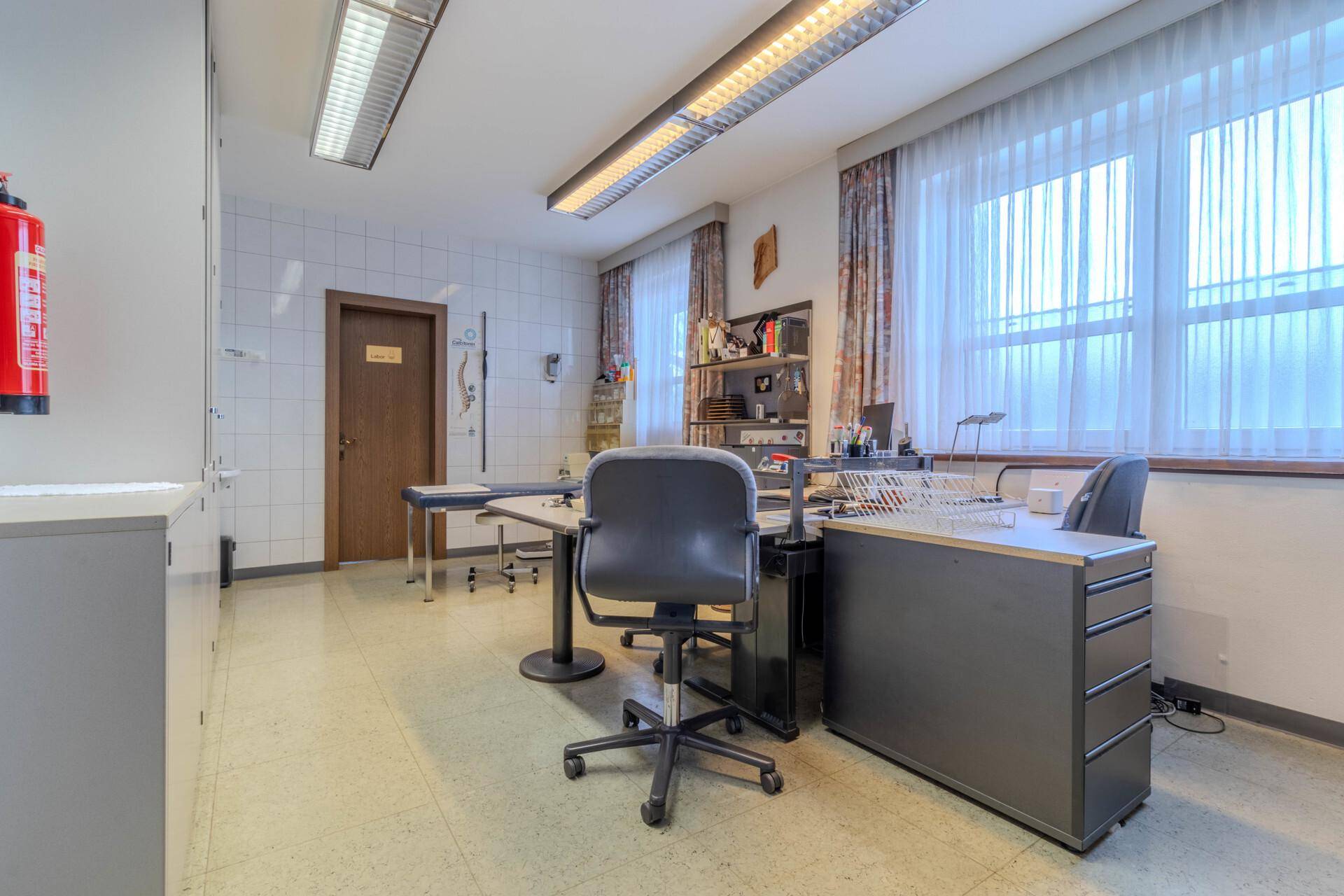 Funktionales Behandlungszimmer mit Schreibtisch, Bürostuhl, Stauraum und großen Fenstern mit Vorhängen.