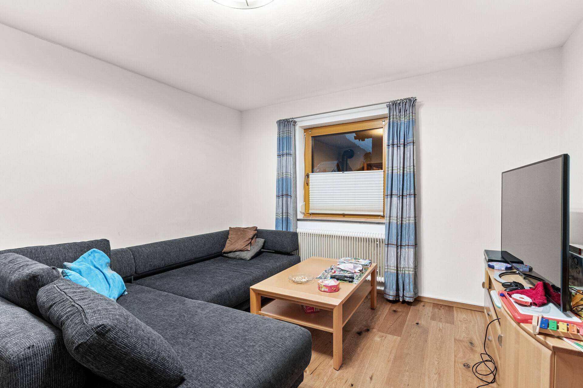 Helles Wohnzimmer mit großem Ecksofa, Holzboden und einem Fenster, das viel Tageslicht hereinlässt.